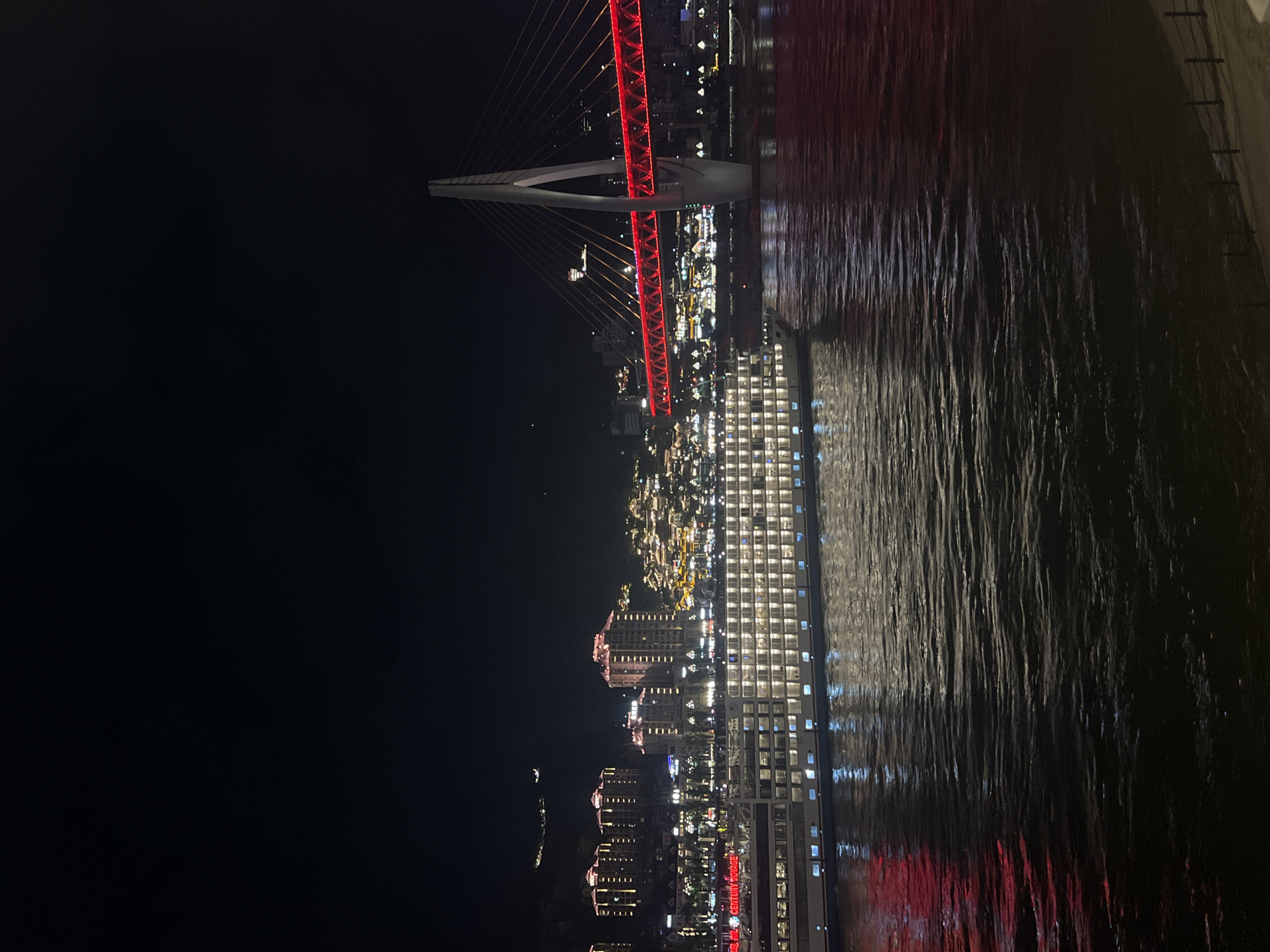 This is a night-time photograph of the Yuzhong District in Chongqing, China. The image features a large, illuminated river cruise ship docked on the Yangtze River. The ship, identified by red lettering on its side as "CENTURY VOYAGE," has multiple decks with numerous lit windows. In the background, a modern suspension bridge with a distinctive arch is prominently displayed. The bridge's deck is illuminated with vibrant red lights, creating a striking contrast against the dark night sky. Behind the bridge, the city skyline rises with illuminated buildings, some of which appear to be hotels or residential towers, displaying warm, yellow lighting. The mountainside behind the buildings is also dotted with lights, suggesting a densely populated urban area. The river's surface reflects the lights of the ship and the bridge, creating a shimmering effect. The overall scene captures the dynamic and illuminated cityscape of Chongqing at night.