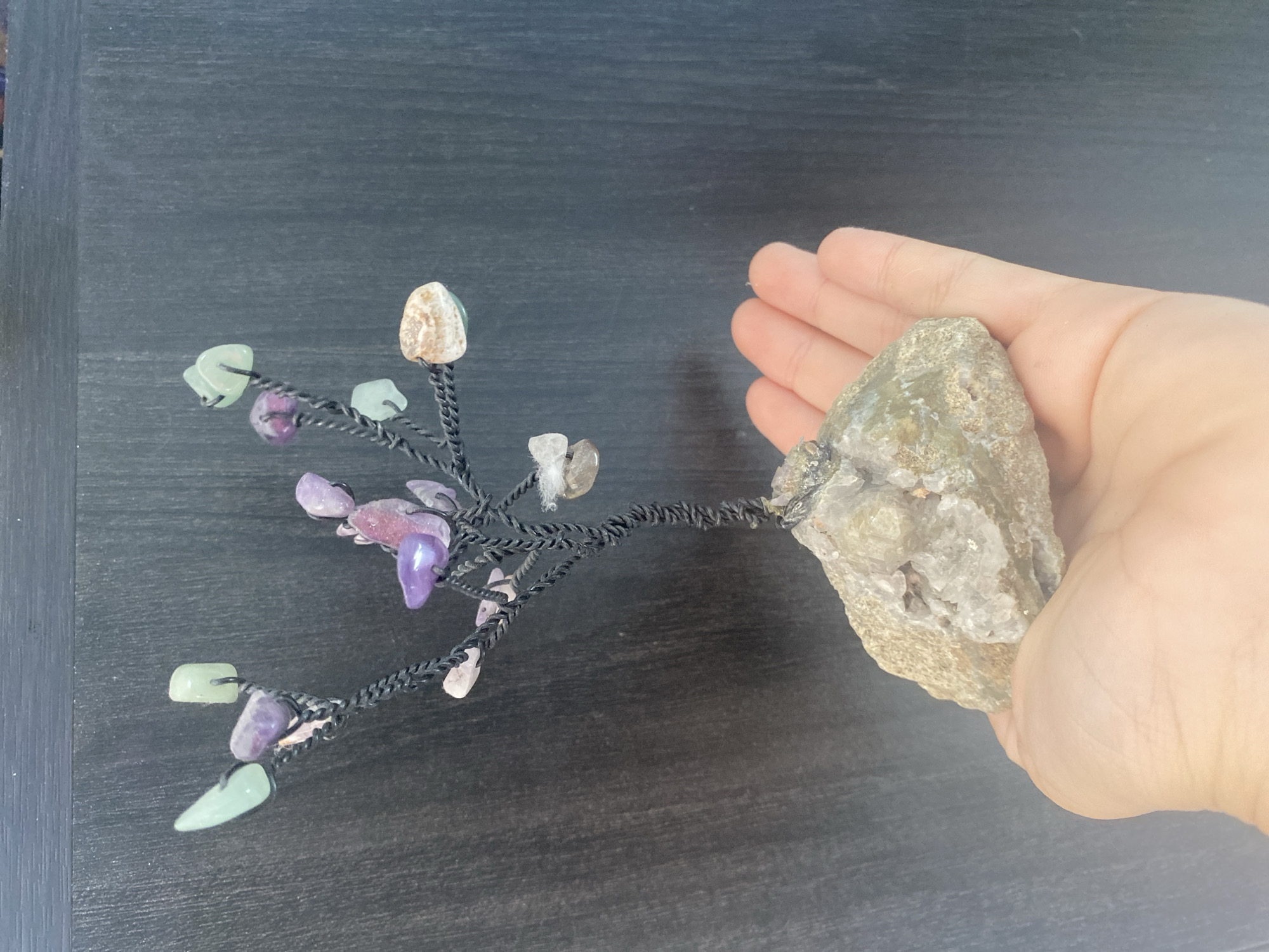 A close-up, eye-level shot shows a hand holding a decorative item. The item is a small, wire-wrapped tree with various polished stones as leaves and fruits, mounted on a rough, light-colored rock base. The stones include shades of purple, green, white, and a peachy-brown, with some appearing faceted. The wire forming the tree is dark and twisted. The hand holding the rock is visible from the palm upwards, with fingers gently supporting the base. The background is a dark, textured surface, possibly a wall or furniture, with vertical lines. The lighting is diffused. There is no visible indication of the city of Blagoevgrad, Bulgaria in the image.