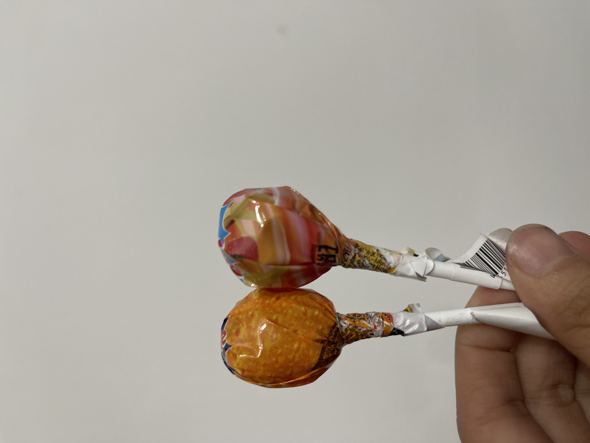 Two lollipops are held by a person's hand against a plain, light-colored background. The lollipops are individually wrapped in colorful cellophane. One lollipop, on the left, has an orange wrapper with a speckled pattern. The other lollipop, on the right, has a wrapper with swirling colors of orange, yellow, pink, and blue, with fruit imagery. The sticks of the lollipops are also wrapped in paper. The background is a uniform, light gray or white, suggesting an indoor setting. The image does not provide any visual information that could be used to determine its location in Bishkek City, Kyrgyzstan.