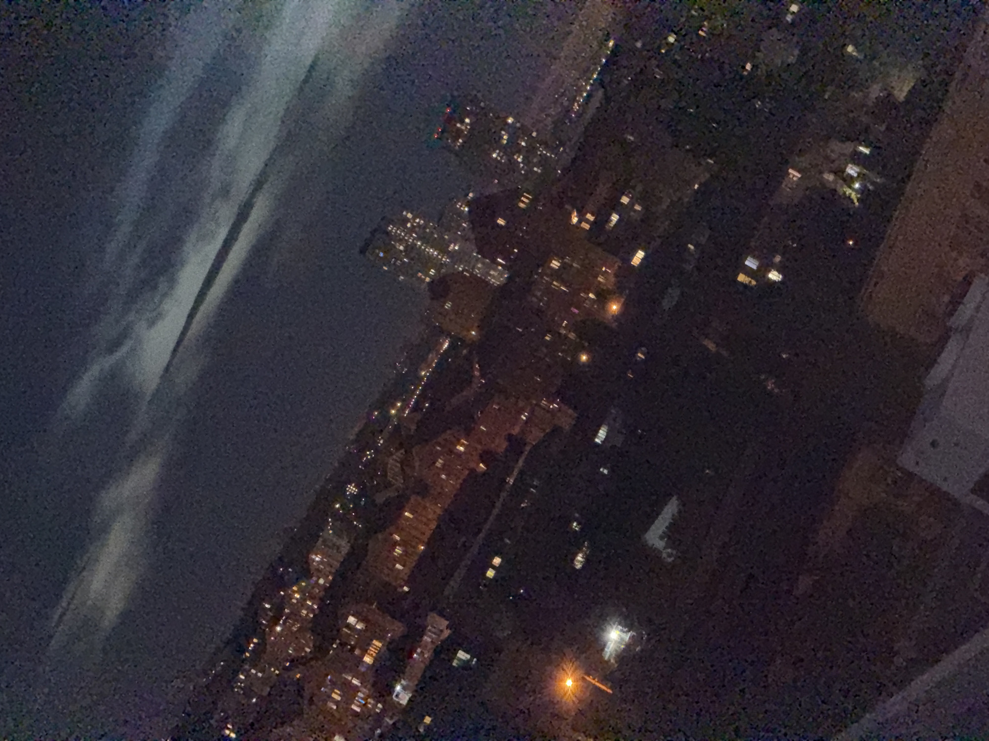 This is a nighttime aerial photograph of an urban landscape, likely a residential area within Kyiv, Ukraine. The image is tilted, giving a diagonal perspective of the scene. The sky above is dark with visible clouds, some illuminated by faint light, suggesting moonlight or city glow.
Below the sky is a densely built-up area with numerous buildings of varying heights. Many of these buildings have illuminated windows, indicating occupancy or active lighting within. The buildings appear to be a mix of apartment complexes and possibly some commercial structures. Some taller, more modern buildings with many windows are visible in the mid-ground and background, characterized by their brightly lit facades. In the foreground and mid-ground, lower-rise buildings are also present, with scattered lights.
A street lamp with a bright orange light is visible on the left side of the image, illuminating a small area below it. The overall impression is of a sprawling city at night, with a distinct urban texture defined by the arrangement of buildings and their lights against the dark sky. The granular quality of the image suggests it was taken with a mobile device in low-light conditions, or possibly at a distance, contributing to a slightly blurred and noisy visual effect. No individuals are discernible in the image.
