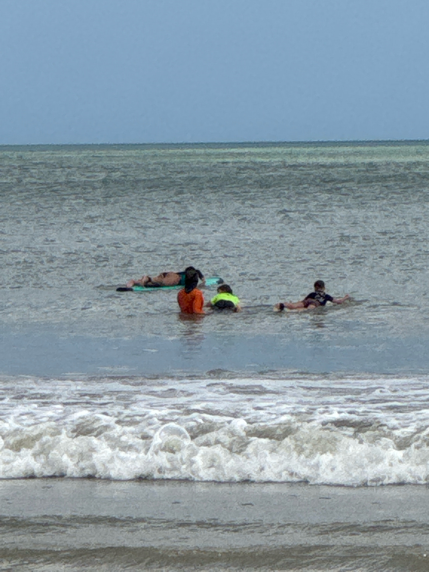 In the ocean off the coast of Kuta Selatan, Indonesia, four people are engaged in watersports. Two individuals are positioned on a green surfboard, one lying prone and the other in a kneeling position. A third person, wearing a bright orange shirt, is partially submerged in the water. A fourth person, wearing a dark shirt and shorts, is also in the water. The sea is calm, with gentle waves approaching the shore. The sky is overcast.