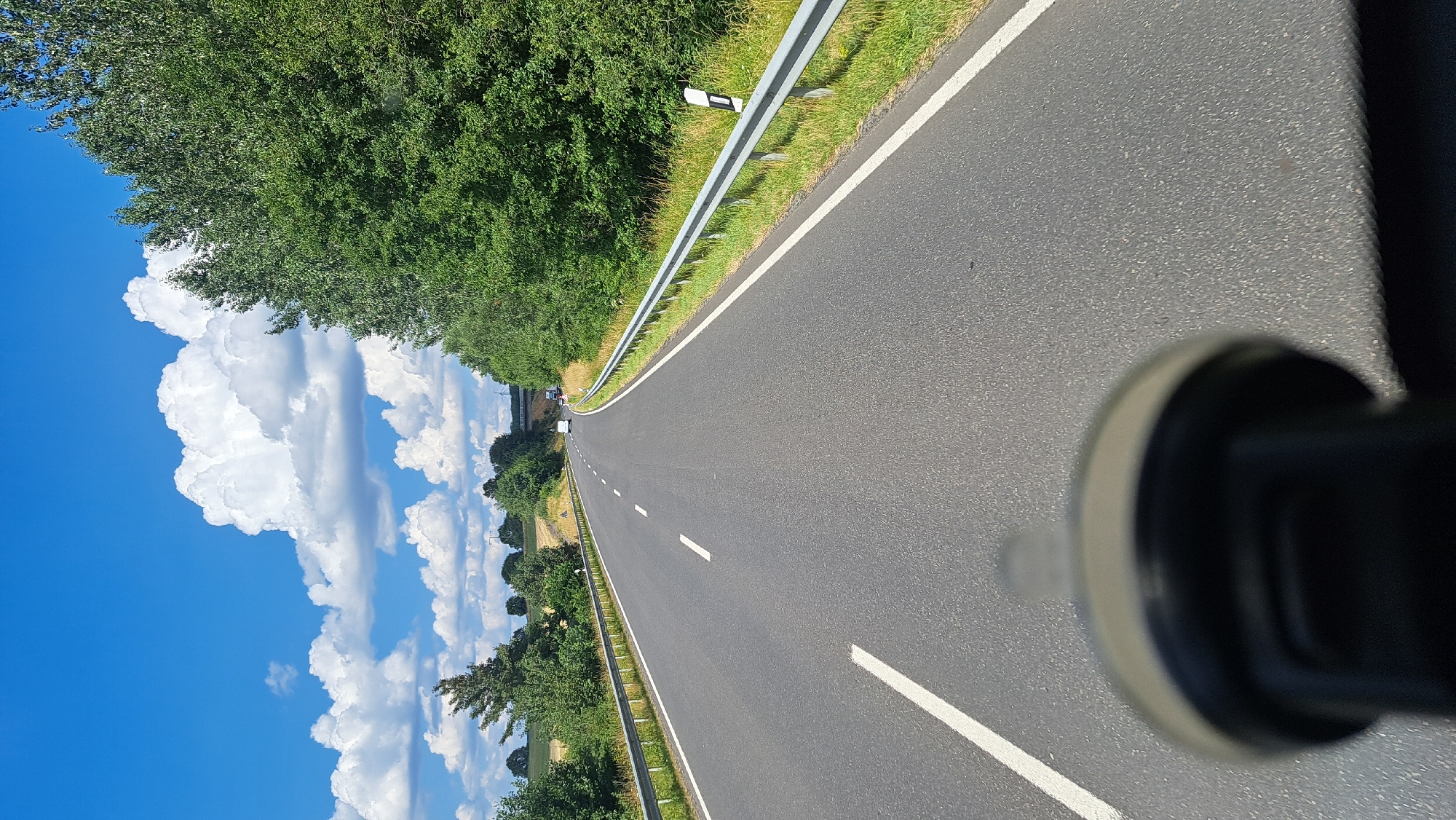 A first-person perspective shot captures a view down a two-lane asphalt road with a white dashed line marking the center and solid white lines along the edges. The road curves gently to the left and disappears over a rise in the distance. To the right of the road, a metal guardrail runs along the edge, bordered by a grassy shoulder and dense green trees. To the left of the road, a similar grassy shoulder transitions to fields and scattered trees. The sky above is a vibrant blue with large, fluffy white cumulus clouds. In the far distance, a white vehicle is visible on the road, and a bridge or overpass is suggested. The foreground is blurred and occupied by a dark, round object, possibly a car mount for a camera or phone. The scene is set in a rural environment, specifically identified as being near Rennerod, Germany. The time of day appears to be midday, given the bright sunlight and clear sky.