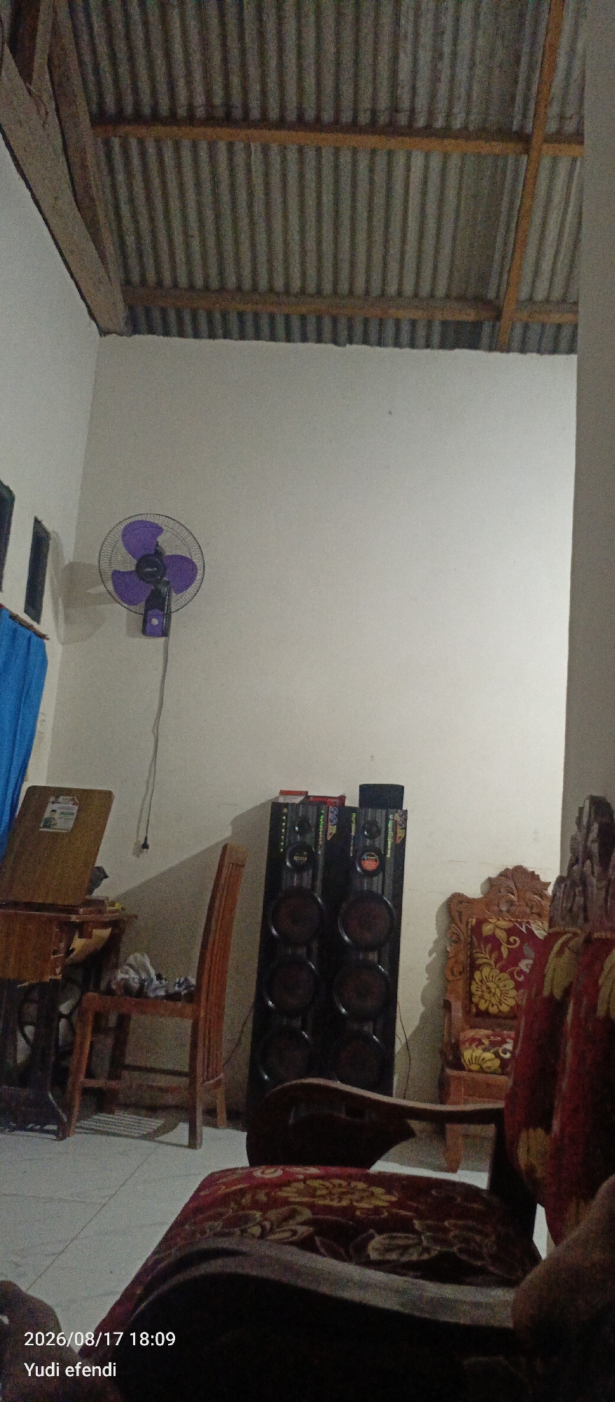 The image depicts an indoor scene, likely a living room or study area, in Rawajitu Selatan, Indonesia. The ceiling is made of corrugated metal panels supported by wooden beams. On the wall, a purple oscillating fan is mounted. To the left, a blue curtain is visible, partially obscuring a window. In the center of the frame, a wooden chair with a patterned cushion stands next to a tall, black speaker system. To the left of the chair and speaker is a wooden desk or table with a slanted surface, possibly an easel or a sewing machine table. Another ornate wooden chair with red and yellow floral upholstery is situated to the right of the speaker. In the foreground, a dark-colored chair with a red and yellow patterned cushion is prominently featured. The floor appears to be tiled. The timestamp "2026/08/17 18:09" and the name "Yudi efendi" are overlaid on the bottom left of the image.