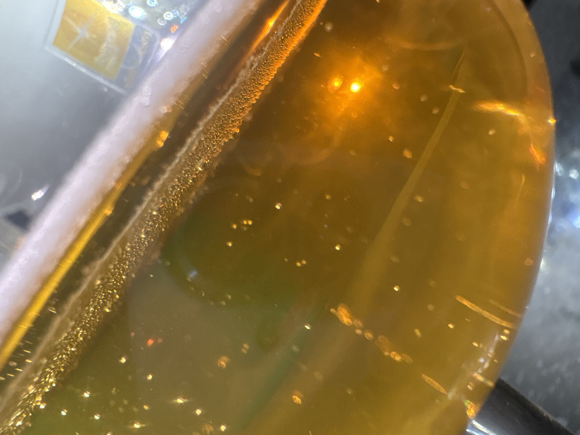 The image displays a close-up view of a liquid, likely a beverage, in a glass container. The liquid is a warm, golden-yellow hue, and appears to contain numerous small bubbles, particularly concentrated along a diagonal line across the upper portion of the frame. These bubbles create a textured, frothy appearance on the surface or within the liquid. Scattered throughout the liquid are small, bright specks of light, suggesting either reflections or suspended particles. The lighting in the scene is dramatic, with strong highlights and areas of deep shadow, giving the liquid a luminous quality. The background is out of focus and appears to be a mixture of light and dark colors, with a hint of a rectangular, possibly labeled, object in the upper right. The angle of the shot is from above and to the side, looking down into the container. There is no discernible information within the image that could link it to the city of Georgiyevsk, Russia.