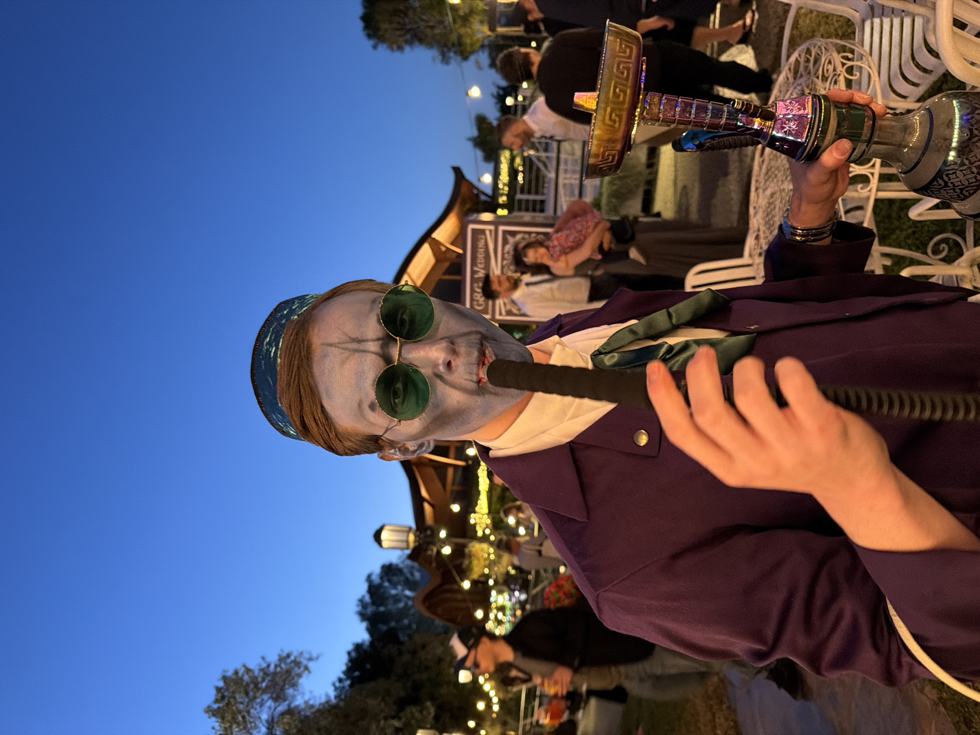This full-shot, portrait-oriented image captures an outdoor evening social event, likely a wedding, as indicated by the visible "GRA WEDDING" sign in the background. The sky is a deep twilight blue, illuminated by numerous string lights and other decorative lighting fixtures that create a festive atmosphere.
The primary subject is a person in a theatrical costume, prominently featured in the foreground. Their face is painted a light blue or grey with dark, skeletal-like contouring under the eyes and on the cheeks. They wear round, green-tinted sunglasses and a patterned blue-green skullcap or head covering. The individual is dressed in a dark purple jacket over a white shirt, with a green tie, and holds an ornate, multi-colored hookah. Their right hand holds the black hose of the hookah to their mouth, while their left hand supports the shimmering base and stem of the hookah, which features metallic gold accents and intricate patterns.
In the blurred background, several casually dressed people are visible, some standing and others seated on white, ornate outdoor chairs around tables. A distinctive wooden structure with a curved roof is also seen behind the main subject. The scene is set outdoors with visible trees and hints of greenery, contributing to a lively and relaxed social gathering under the evening sky.