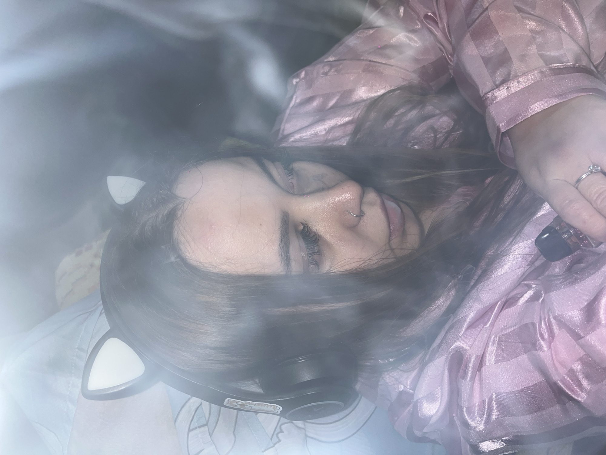 A woman wearing cat-ear headphones and a pink satin pajama set is lying down. She has dark hair and a nose ring. She is also wearing a ring on her right ring finger. The scene is obscured by a hazy or smoky effect. The location in the city of Novopokrovskaya, Russia, is not discernable from the image.