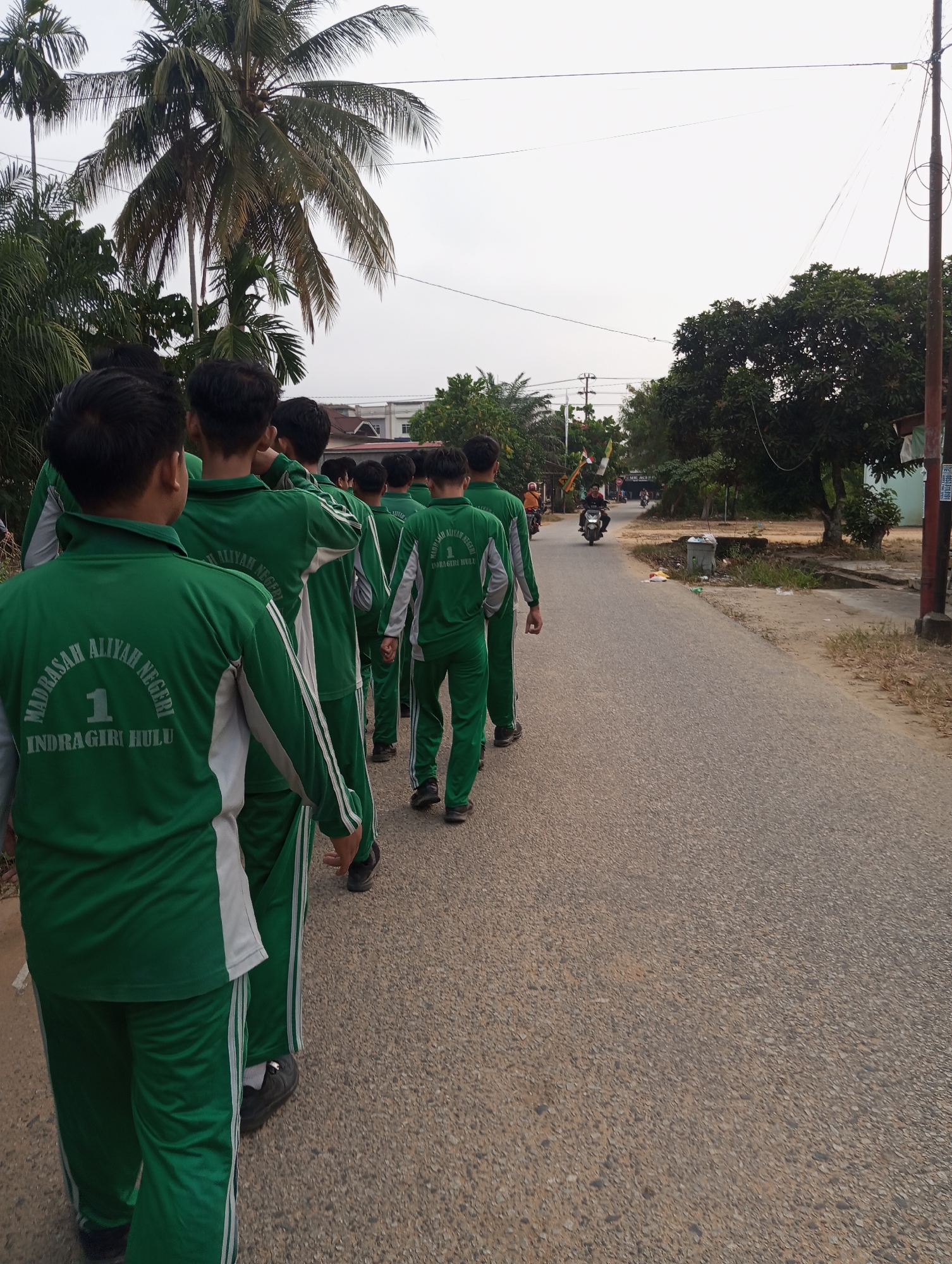 A group of male students, dressed in matching green tracksuits with white accents and the emblem "MADRASAH ALIYAH NEGERI 1 INDRAGIRI HULU," walk in single file along a paved road. The road appears to be a local street in Pematang Reba, Indonesia, indicated by the presence of palm trees and tropical vegetation, as well as a building visible in the background. The students are walking away from the camera, their backs to the viewer. The road is bordered by a grassy shoulder and some scattered debris. In the background, a motorbike and a few other individuals can be seen. The sky is overcast, suggesting it might be early morning or late afternoon. The overall atmosphere is mundane, capturing a common scene of students commuting or participating in an outdoor activity.