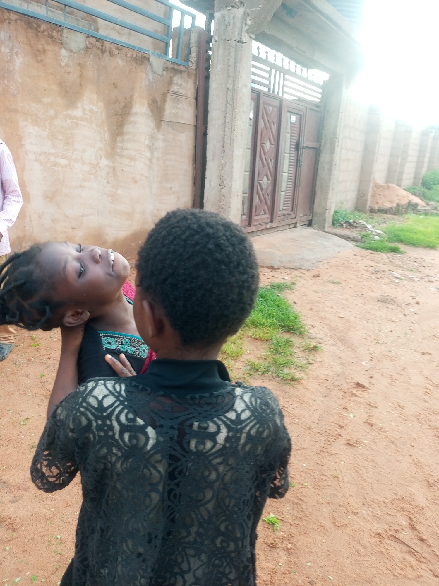 This media file depicts two young Black children interacting outdoors in a village setting in Tempure, Nigeria. The child in the foreground, viewed from the back, has short, dark, curly hair and is wearing a black top with a lace overlay. They are holding a second child, who is facing away from the camera, looking upwards and to the left. This second child has dark braided hair and is wearing a black top with a green and red embroidered neckline.
The ground is sandy, with patches of green grass scattered around. In the background, there is a textured, light brown wall with a metal railing above it. To the right, a large, dark wooden gate with a patterned design is visible, set within a concrete structure that appears to be a doorway or entrance. Further to the right, a brick wall and a pile of sand suggest ongoing construction or building work. The overall lighting suggests it is daytime, possibly late afternoon, with the sun casting a bright glare in the upper right portion of the image. The expression on the face of the child looking upwards seems joyful, with a wide smile and eyes partially closed. The scene appears informal and domestic. No visible text is present in the media file.