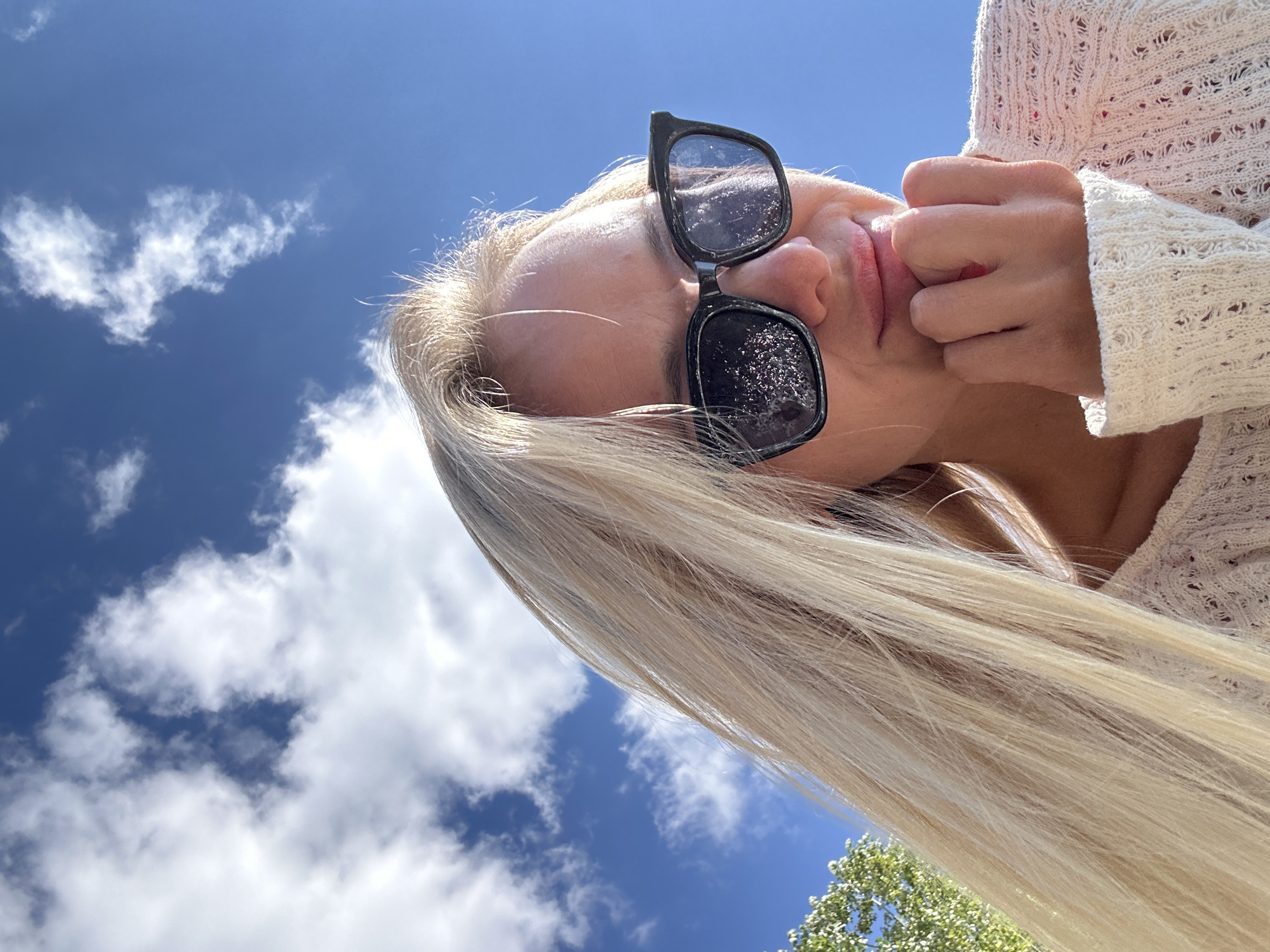A young woman with blonde hair and sunglasses poses for a selfie outdoors, with a bright blue sky and scattered clouds as the background. She is wearing a white, knitted sweater and resting her chin on her hand. The image is likely taken on a sunny day. The location of Gornyi Erik, Russia is not discernible from the image.