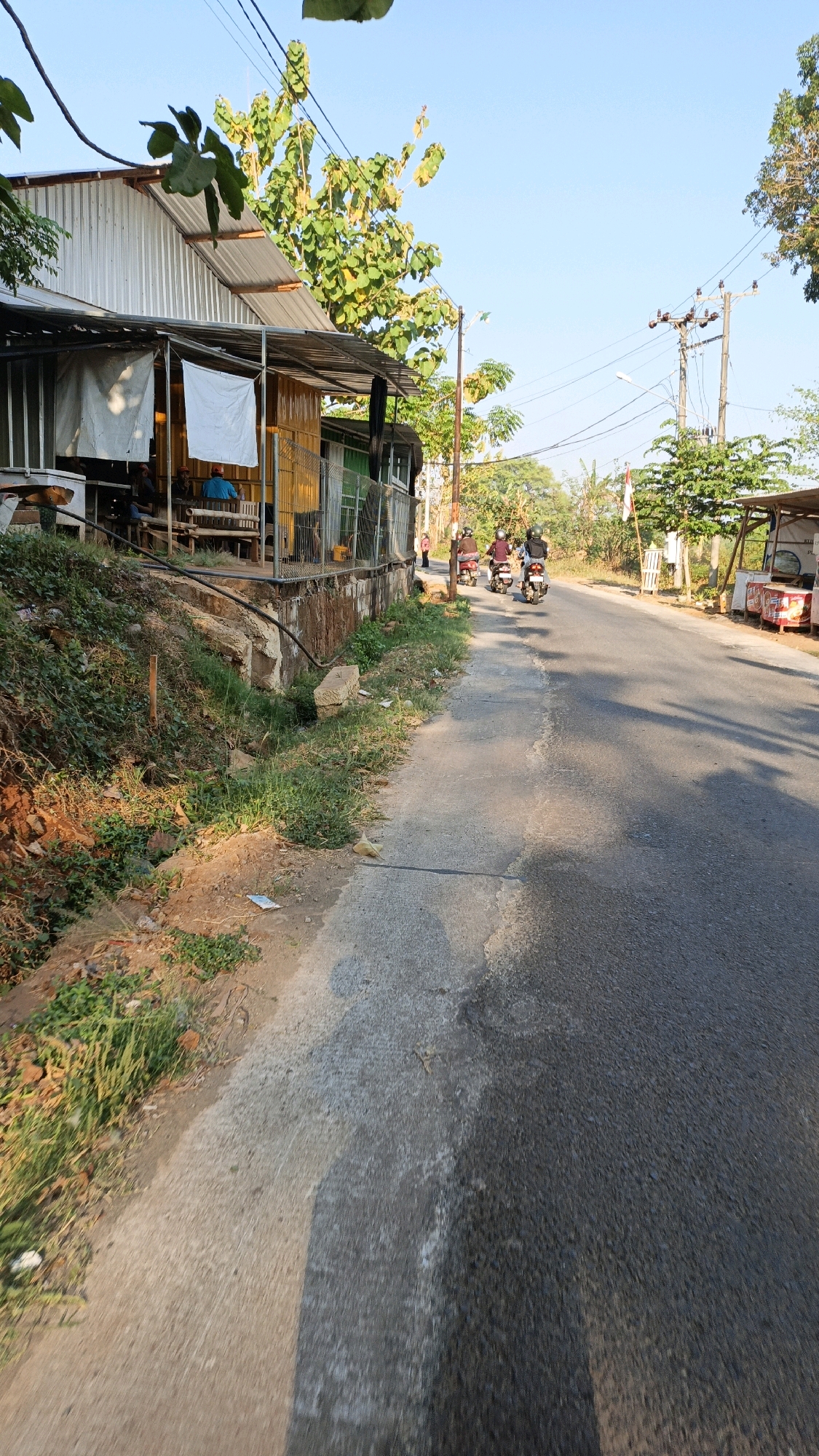 A street-side view in Kuwasen, Indonesia, shows a scene captured during daylight. To the left, a simple, elongated structure with a corrugated metal roof and bamboo walls sits on an elevated embankment. White tarpaulins are hung on the front of the structure, and several people are visible sitting or standing within. A narrow paved path runs alongside the embankment, sloping down to the road. The embankment itself is covered in overgrown grass and dirt, with some visible debris.
The road, paved with asphalt, occupies the right side of the frame and curves gently into the distance. Several motorcycles with riders are traveling down the road away from the viewer. The environment is rural, with trees and vegetation visible on either side of the road. Electrical poles and wires are present along the roadside. The sky is clear and blue, indicating fair weather. The overall impression is of a typical roadside scene in a developing area.