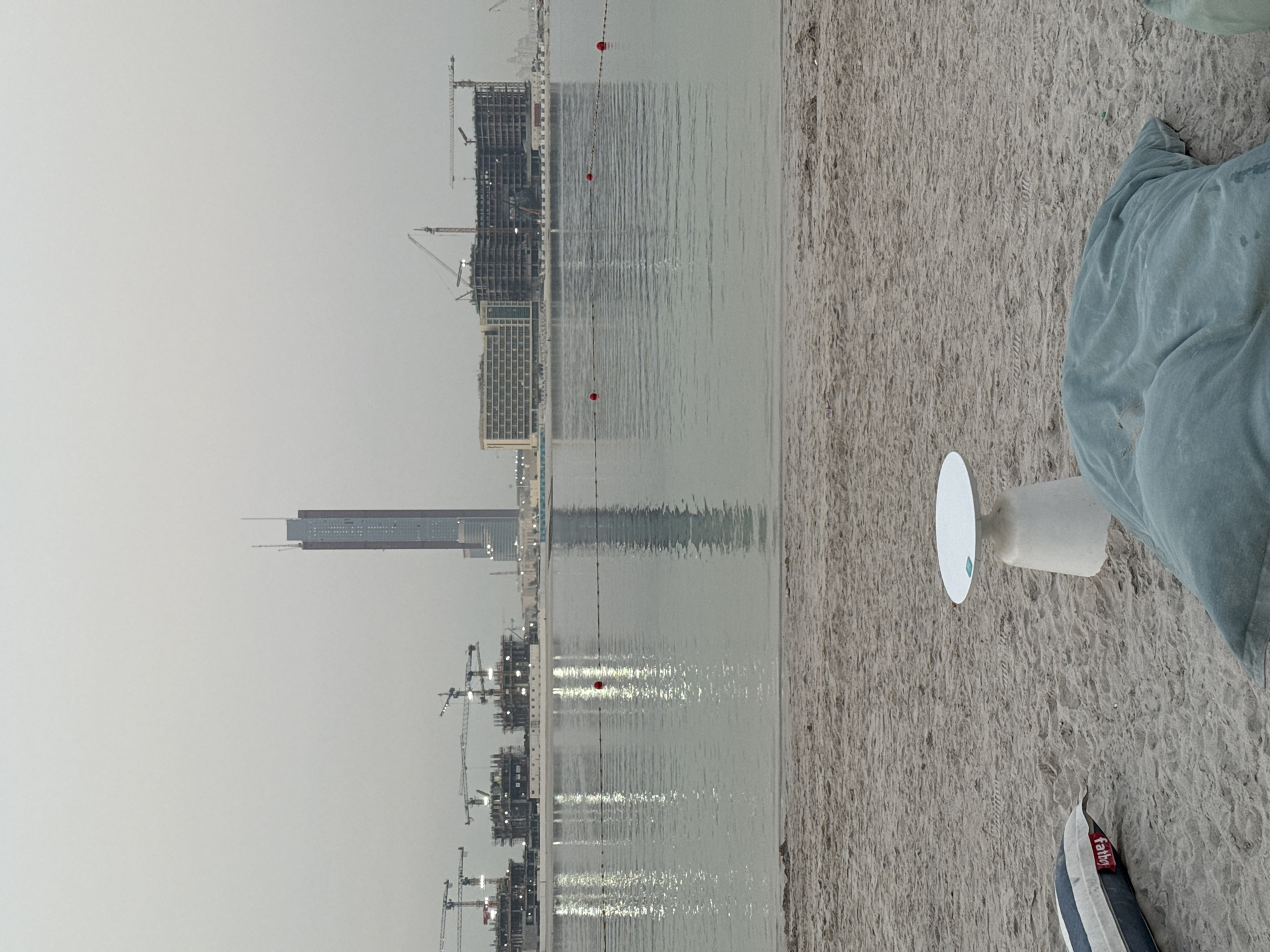 A beachfront scene is depicted with a calm body of water in the mid-ground and a sandy beach in the foreground. The water reflects the light from a string of buoys with red markers. In the background, a city skyline is visible, characterized by construction cranes and several buildings, including a tall, slender skyscraper. The sky is overcast and hazy. On the beach, there are a few pieces of outdoor furniture, including a small white table and large, soft seating cushions. One cushion appears to be a light blue beanbag, and another, partially visible, is blue and white striped. The overall atmosphere suggests a potentially developed but still under-construction area.