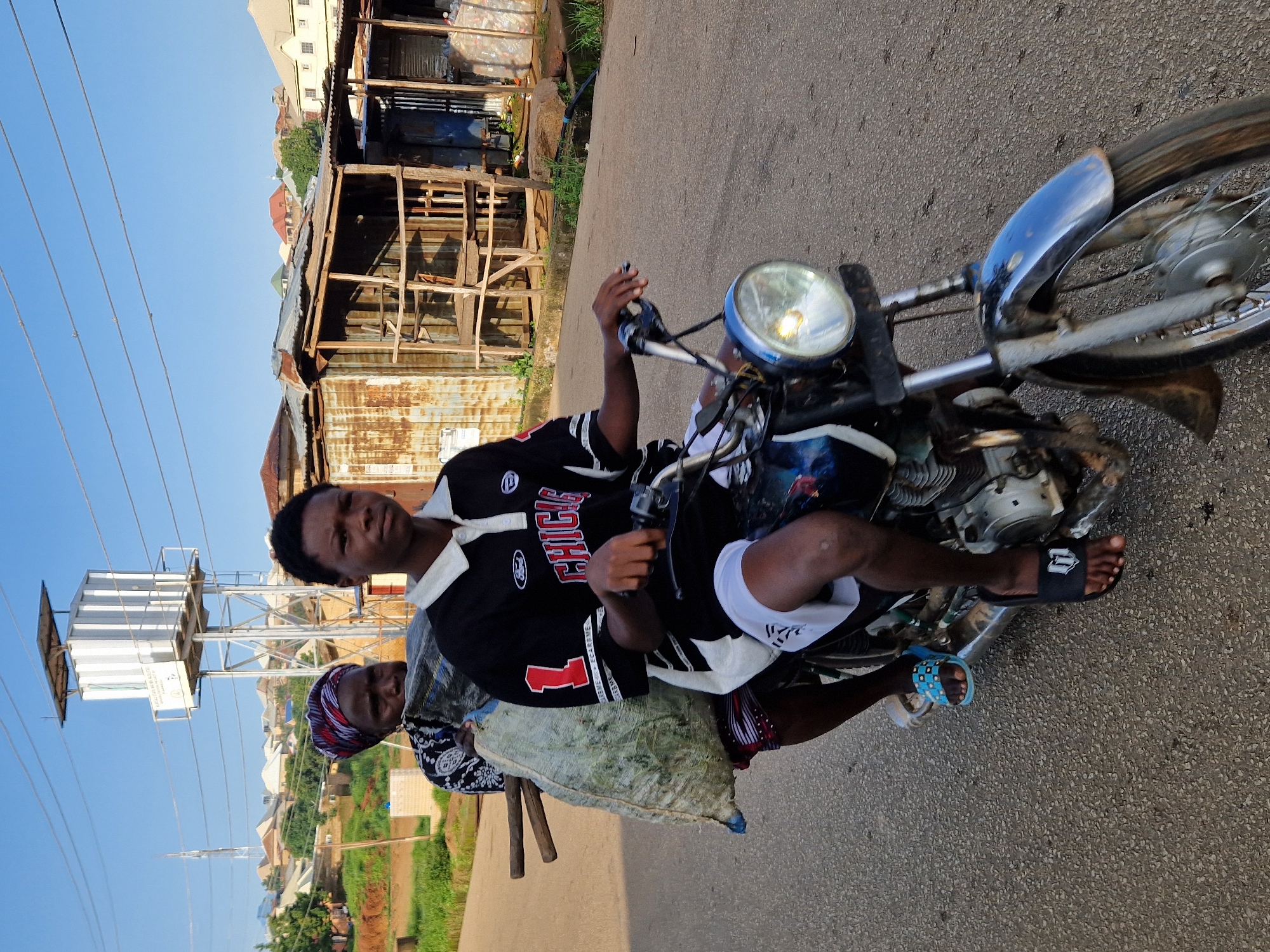 Two individuals are riding on a motorcycle on a paved road. The person in the front is a young male, the driver, wearing a black and white collared shirt and white shorts. The person behind is an older female, wearing a patterned headwrap and a dark and white patterned top. She is holding a large, light-colored bag that is strapped to her back, and two wooden sticks are protruding from the top of the bag. The motorcycle is a dirt bike style with a prominent headlight. The background shows a residential area with a water tower, some low-rise buildings, and vegetation. The sky is clear and blue. The lighting suggests it is daytime.
