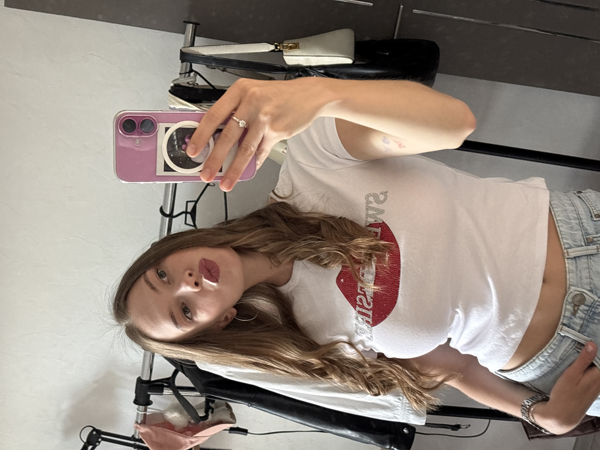 A young woman with long, wavy brown hair is taking a selfie in what appears to be a closet or dressing area. She is wearing a white t-shirt with a large red sequined lip graphic and the word "SWEET" spelled out in rhinestones below it. She is also wearing light-wash denim jeans. Her left hand is holding her phone, which has a pink case and is decorated with a circular sticker. She is wearing a ring on her right hand, which is resting on her chin. In the background, there are several clothing items hanging on a rack, including a white jacket and a black leather jacket. There are also two bags visible, one black and one cream-colored. The environment appears to be indoors, with a plain white wall behind the clothing rack. There are no discernible landmarks or signs that would indicate a specific location within the city of Perm, Russia.