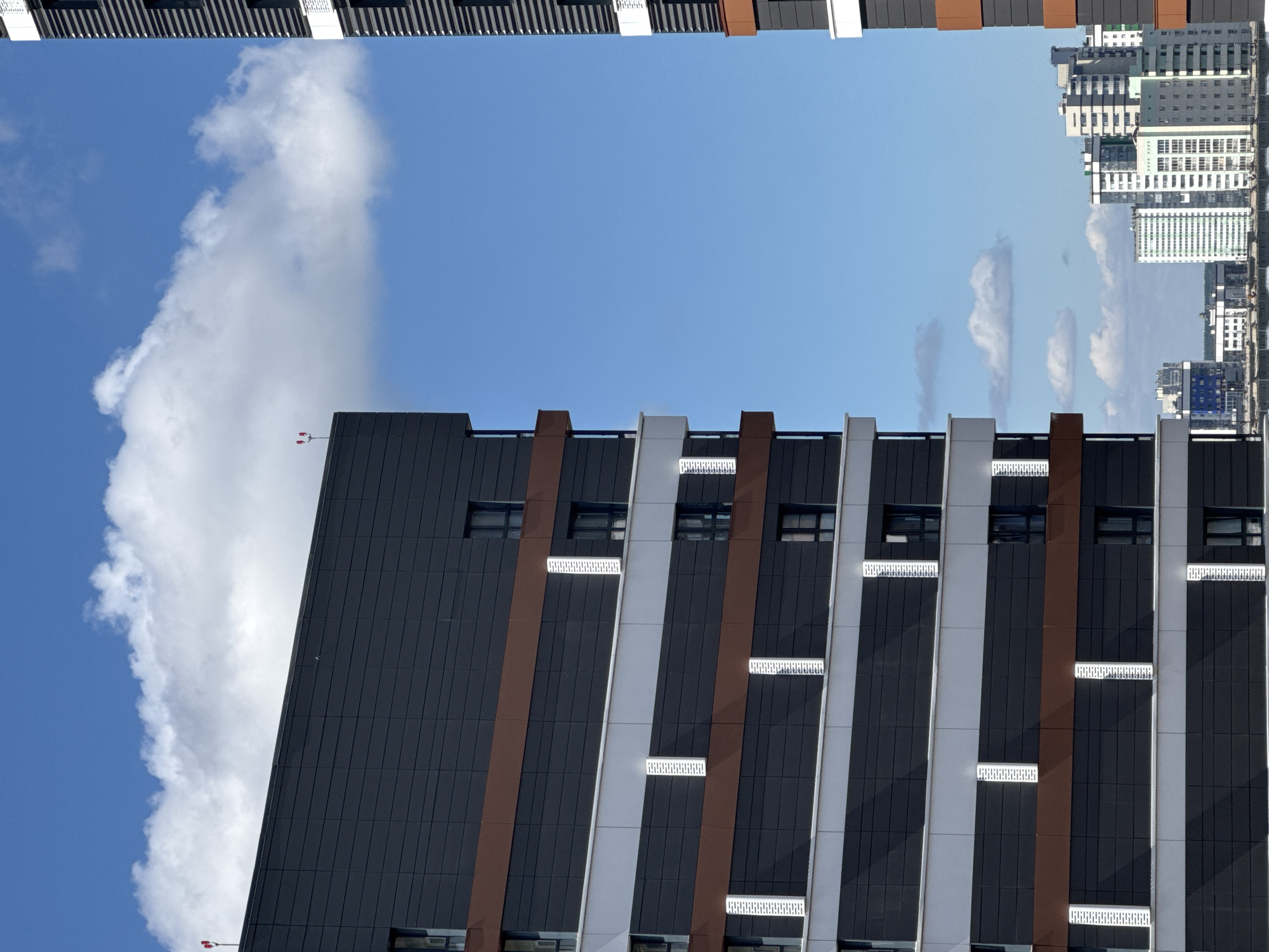 The image shows a partial view of modern, multi-story buildings against a bright blue sky with scattered white clouds. On the left, a dark gray building with horizontal bands of brown and white paneling is prominently featured. Several windows are visible on this building. To the right, another similar building is partially visible, also with dark gray, brown, and white sections, and a striped pattern on its facade. In the background, a cluster of other, more distant buildings of varying architectural styles can be seen, suggesting an urban setting. The scene is likely a daylight shot, possibly taken from a high vantage point overlooking a city. There is no indication of specific people, actions, or interactions. Based on the provided context, this scene is located in the city of Novosibirsk, Russia.