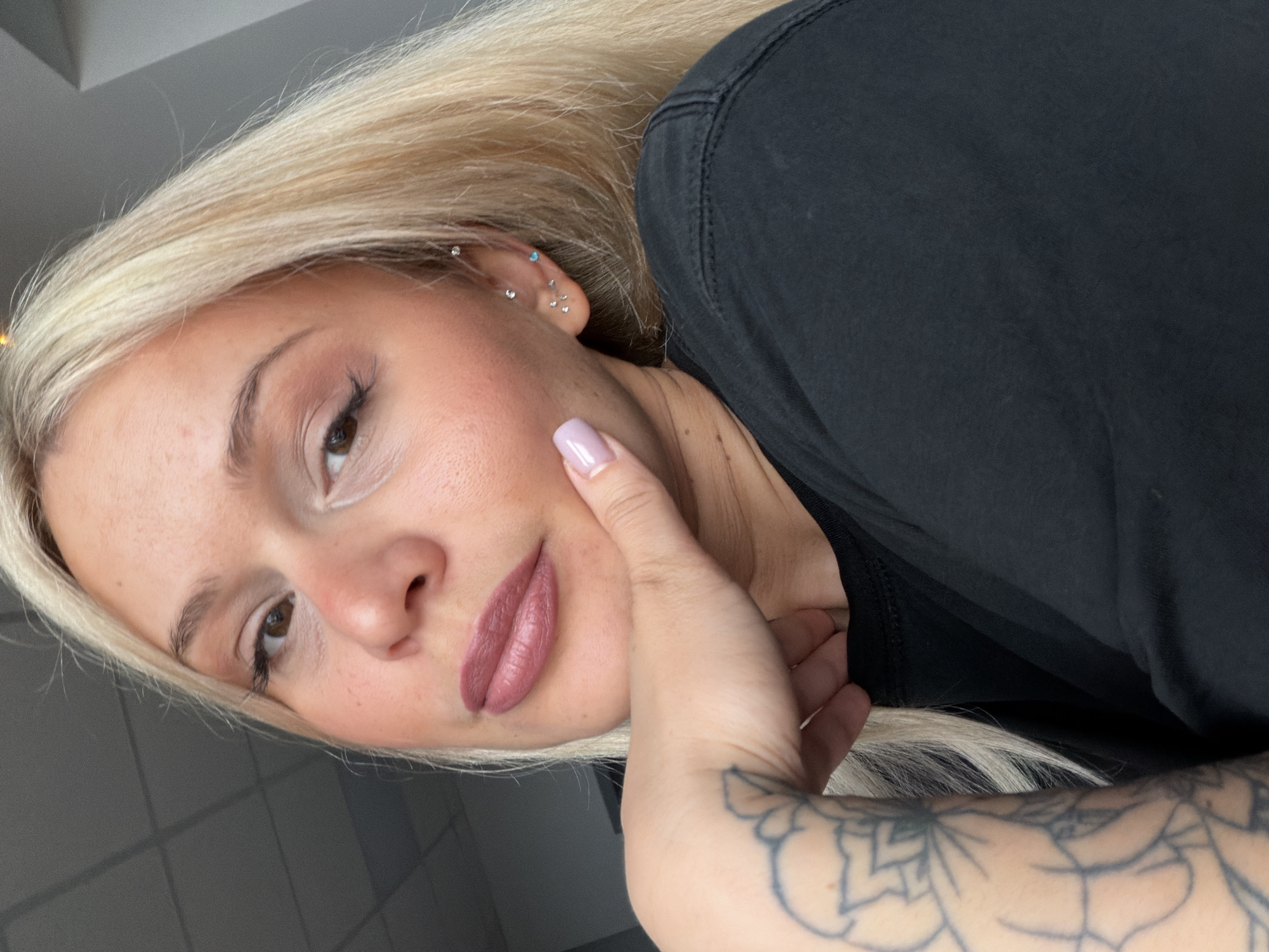 The portrait, likely taken indoors in Sarapul, Russia, captures a young woman with long, straight platinum blonde hair, looking slightly upward and to her right with a neutral and serene expression. Her left hand is gently placed against her jawline, revealing light lavender nail polish on her index finger.
The woman has light brown eyes, well-defined eyebrows, and long eyelashes, enhanced with subtle white eyeliner in the inner corner and a matte mauvy-pink lipstick. Her visible left ear features multiple cartilage piercings, including several small silver-toned studs and one small, light blue stud.
She is dressed in a plain black t-shirt or top. A prominent, intricate tattoo with a floral or mandala-like design is visible on her inner left forearm. The background is blurred and minimalist, displaying muted grey tones and indistinct rectangular patterns, characteristic of an indoor setting. The soft, natural-looking light illuminates her face. There is no visible text in the image.