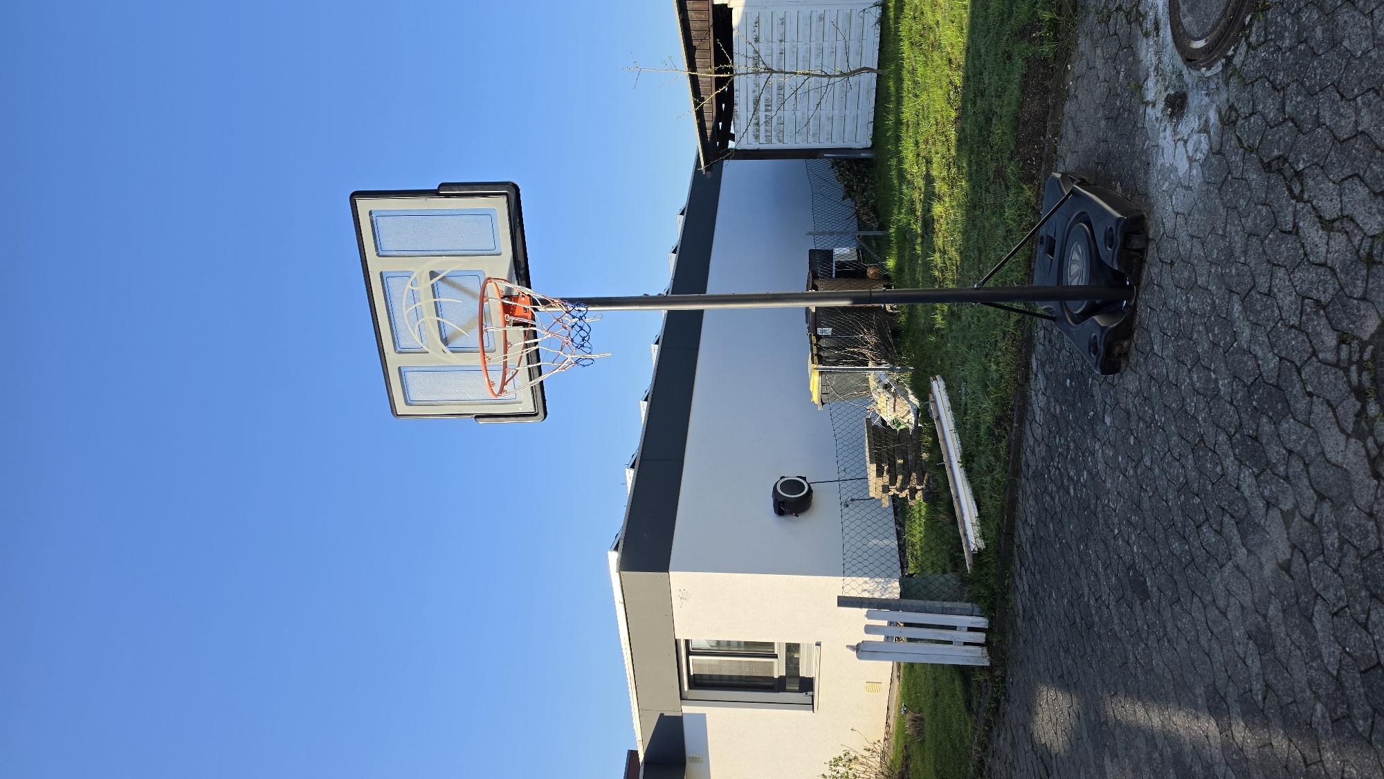 The image depicts a basketball hoop in front of a white building with a gray roof. The hoop has a clear backboard, an orange rim, and a blue and white net. The base of the hoop is black and sits on a paved surface. In the background, there is a green lawn, a white fence, and a white shed. The sky is clear and blue. The location is in the city of Unknown, Unknown.