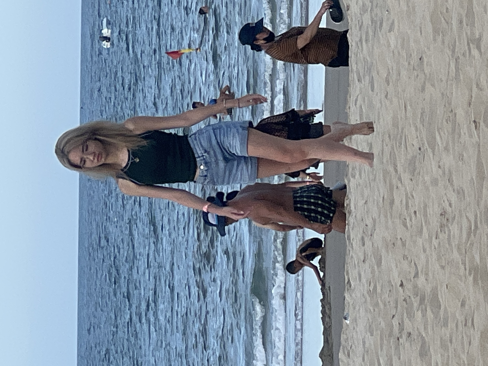 The image depicts a beach scene in Da Nang, Vietnam, with the ocean in the background and sand in the foreground. A young woman with blonde hair, wearing a dark green tank top and denim shorts, is walking towards the viewer in the center of the frame. Several other people are visible on the beach and in the water. To the left, a child is playing in the sand, and a man wearing checkered shorts and a hat is standing. In the background, people are swimming in the ocean, and a small boat is visible on the water. A red and yellow flag, likely a lifeguard marker, is planted in the sand near the water's edge. The overall atmosphere appears relaxed and casual, characteristic of a beach resort area.