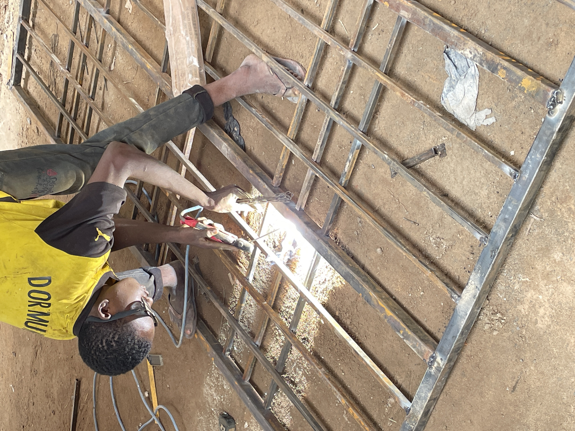 In Okinano, Nigeria, a young man is seen welding a metal gate outdoors. He is wearing a yellow t-shirt with black sleeves and black pants, and he has safety glasses on. The welding is taking place on a flat, dirt surface, which is typical of many outdoor work areas in the region. The metal gate is laid out on the ground, and the man is working diligently to join the metal bars together. Sparks are flying from the welding torch, illuminating the immediate area and highlighting the intense heat of the process. The surrounding environment appears to be a workshop or a construction site, with other metal materials and tools potentially present but not clearly visible in the frame. The overall scene depicts a craftsman at work, contributing to the infrastructure or livelihood of the community.