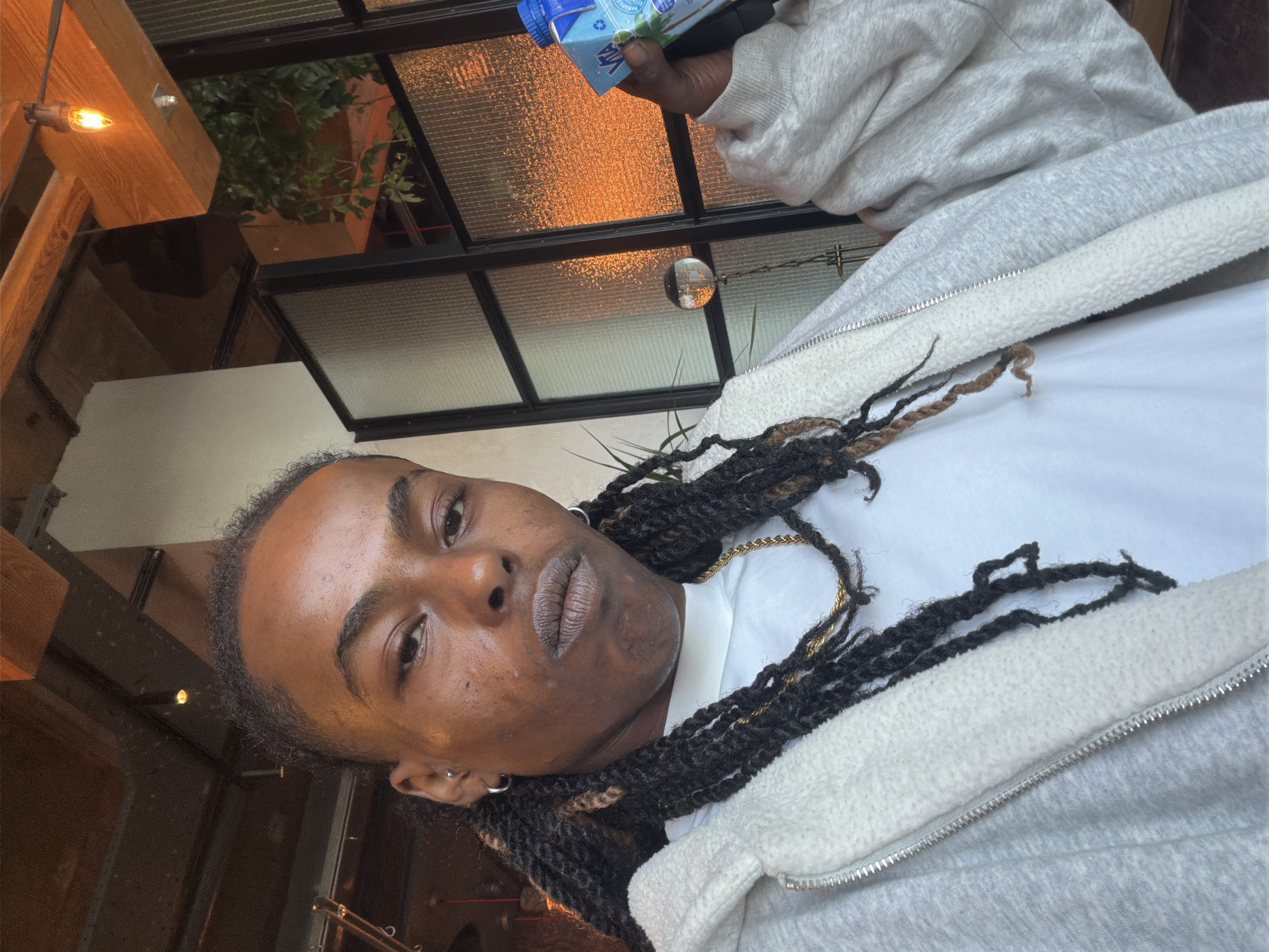 The image captures a person, appearing to be male, with dark skin and short, twisted dreadlocks that include black and brown strands. He looks directly at the camera with a neutral expression. He is wearing a white crew-neck t-shirt and a light grey zipped hoodie, which features a fluffy, sherpa-like lining visible around the opening. A gold chain is visible around his neck, and he has small hoop earrings in both earlobes, with an additional stud in the tragus of his left ear.
In his right hand, the person holds a small, light blue carton. Visible text on the carton includes "VITA" and "COCONUT WATER."
The setting is indoors, likely a cafe or restaurant in London, characterized by modern, industrial-style architecture. The background features a grid of windows with black frames and textured or frosted glass panes, some reflecting a bright, circular light source. Warm, artificial light, possibly from exposed bulbs, casts an orange glow across parts of the ceiling and some window surfaces, suggesting an evening ambiance or a richly lit interior. Green plants are visible through some of the window sections, hinting at either an outdoor view or indoor greenery. Wooden beams or structural elements are also visible in the upper left corner of the frame. The overall perspective suggests the person is leaning back or reclining, with the scene appearing slightly rotated.