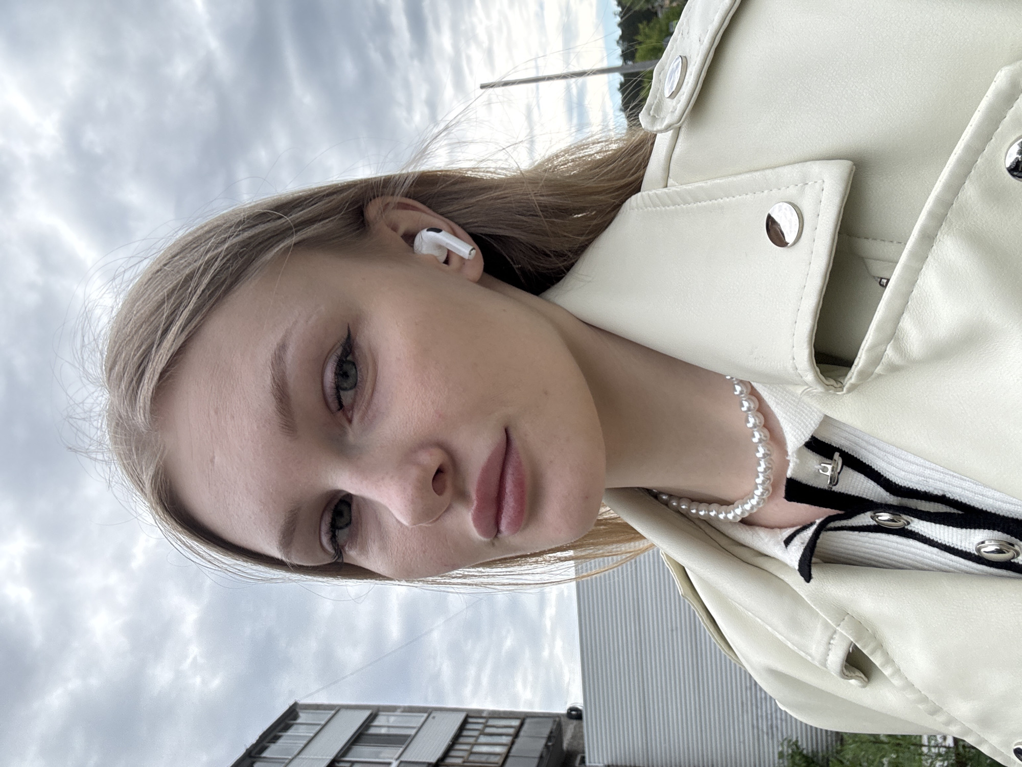 A young woman, appearing to be in her late teens or early twenties, is depicted in a close-up, chest-up outdoor portrait taken in Perm, Russia. She has long, straight, light-blonde hair, green eyes with dark winged eyeliner, and full, light pink lips. Her expression is a subtle, direct gaze towards the camera. She wears a cream or off-white faux leather jacket with prominent silver snap buttons on the lapels and shoulder tabs. Beneath the jacket, a white ribbed top or cardigan with black horizontal stripes and a silver hook clasp is visible. A single strand of white pearls adorns her neck. A white Apple AirPod Pro is inserted into her right ear. The background consists of a heavily overcast, grey sky. To the left, a portion of a multi-story residential building with dark windows is visible. A light grey corrugated fence or wall is situated behind her lower left side. On the right, a tall, thin, dark pole stands against a backdrop of distant green foliage.