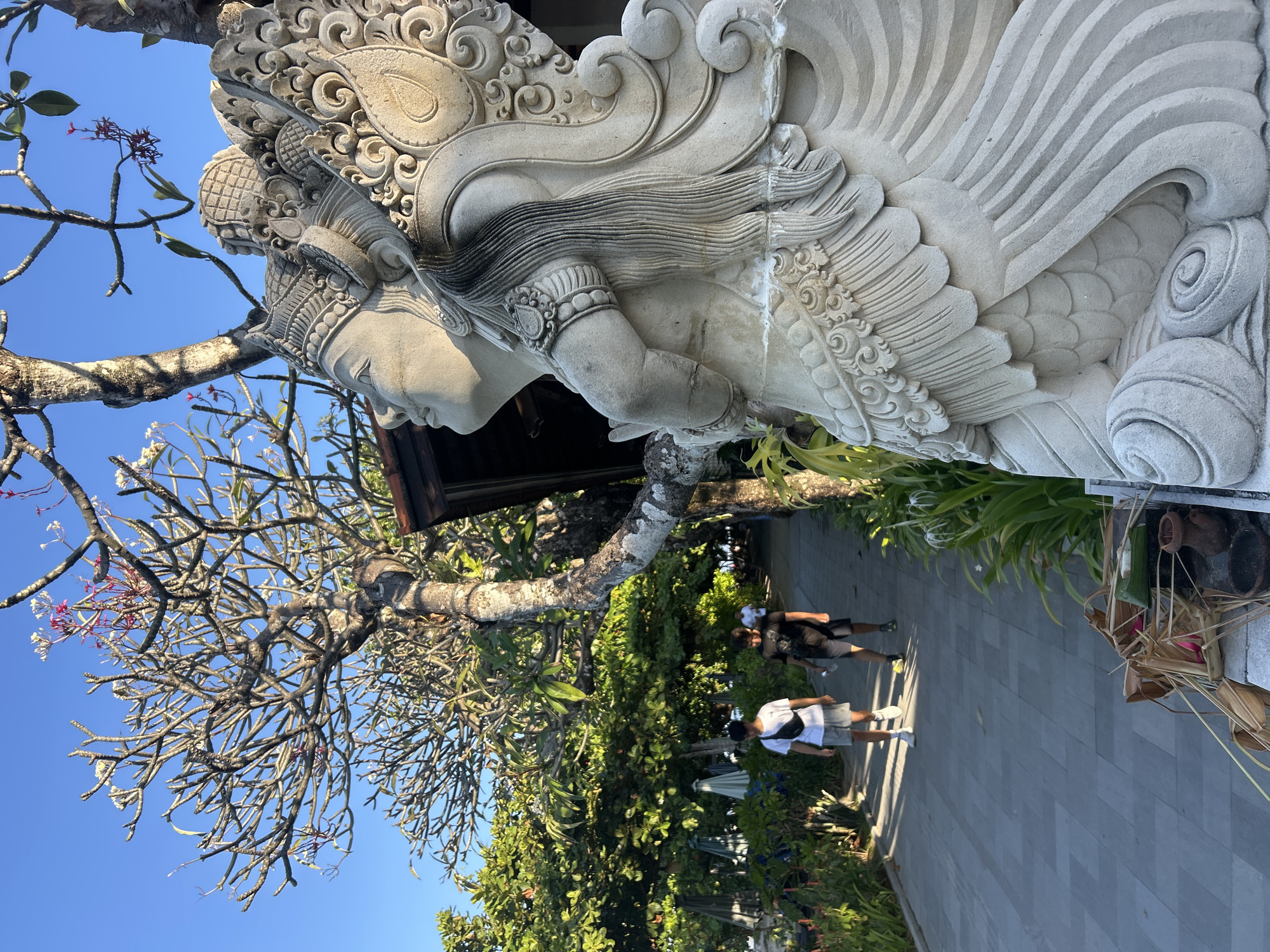 In Denpasar, Indonesia, a stone statue of a deity with elaborate headdress and feathered wings stands prominently in the foreground on the right. The statue is positioned next to a pathway made of grey paving stones. Two young men are walking down the path towards the viewer. The man on the left wears a white t-shirt, grey shorts, and white sneakers, with a black fanny pack slung across his chest. The man on the right wears a black t-shirt, dark shorts, and dark athletic shoes, carrying a water bottle. Behind them, lush green trees and vegetation line the path, with some beach umbrellas visible in the distance. The sky is clear blue. The statue's weathered appearance suggests it is an outdoor sculpture, likely in a garden or temple setting. A small offering of food and some straw bundles are placed at the base of the statue.