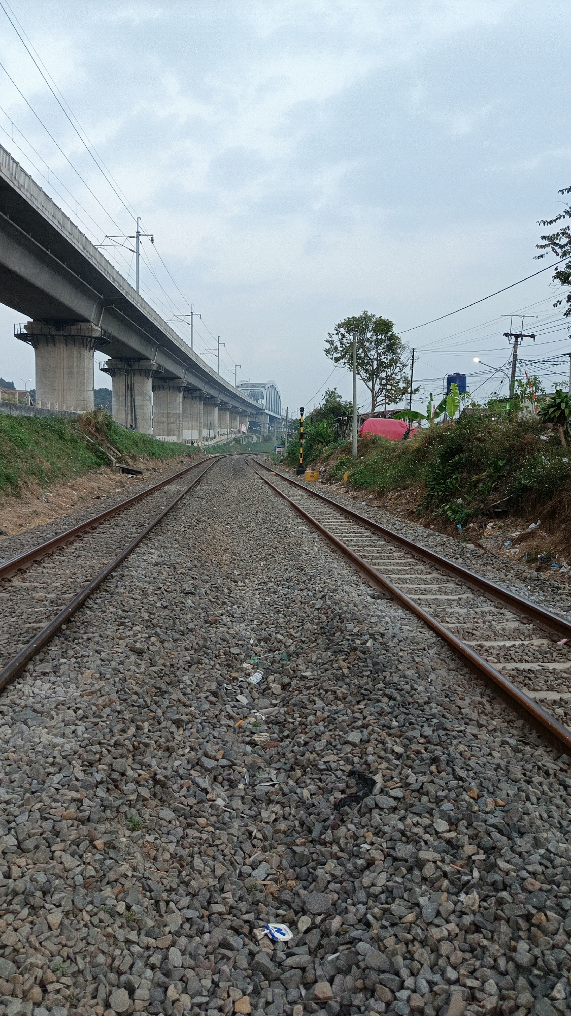 The image captures a pair of railway tracks diverging into the distance, with a concrete elevated structure, likely a highway or a metro line, running parallel to the tracks on the left. The railway bed is composed of grey ballast stones. In the background, on the right side of the tracks, there is vegetation, a red tarp, and utility poles with wires. A yellow and black striped pole, possibly a signal, is visible near the right track. The sky is overcast with clouds. The scene appears to be located in a semi-urban or peri-urban area of Margajaya, Indonesia, characterized by infrastructure development and surrounding greenery.