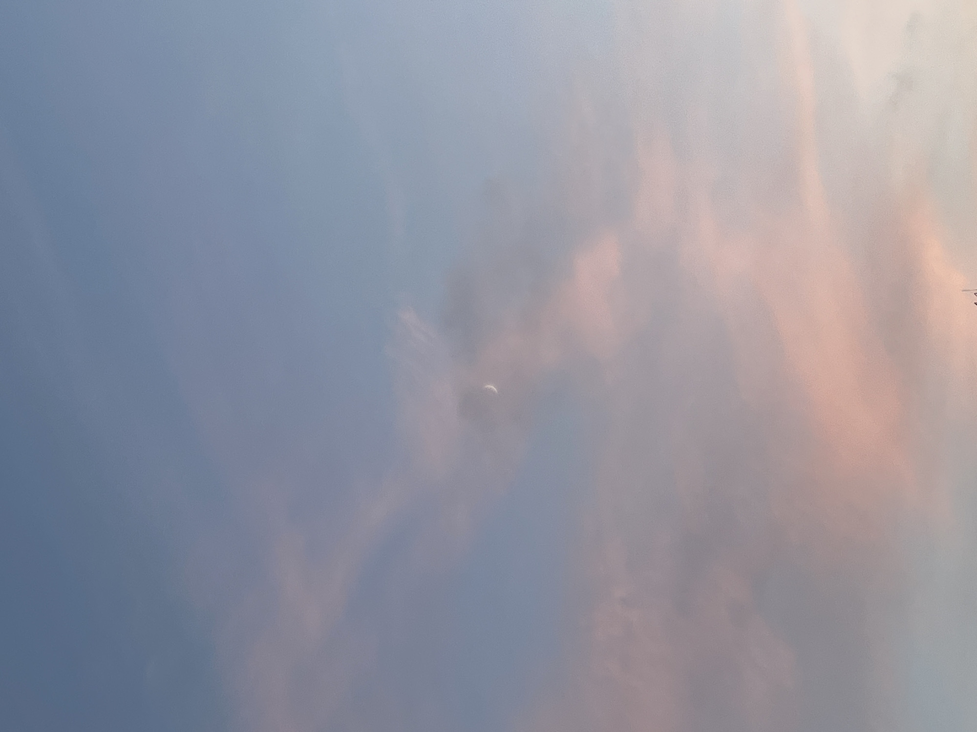 The image shows a daytime sky in Saint Petersburg, Russia, featuring a thin crescent moon partially obscured by wispy clouds. The sky has a gradient of light blue to pink hues, suggestive of either sunrise or sunset. There are no people or discernible objects in the foreground.