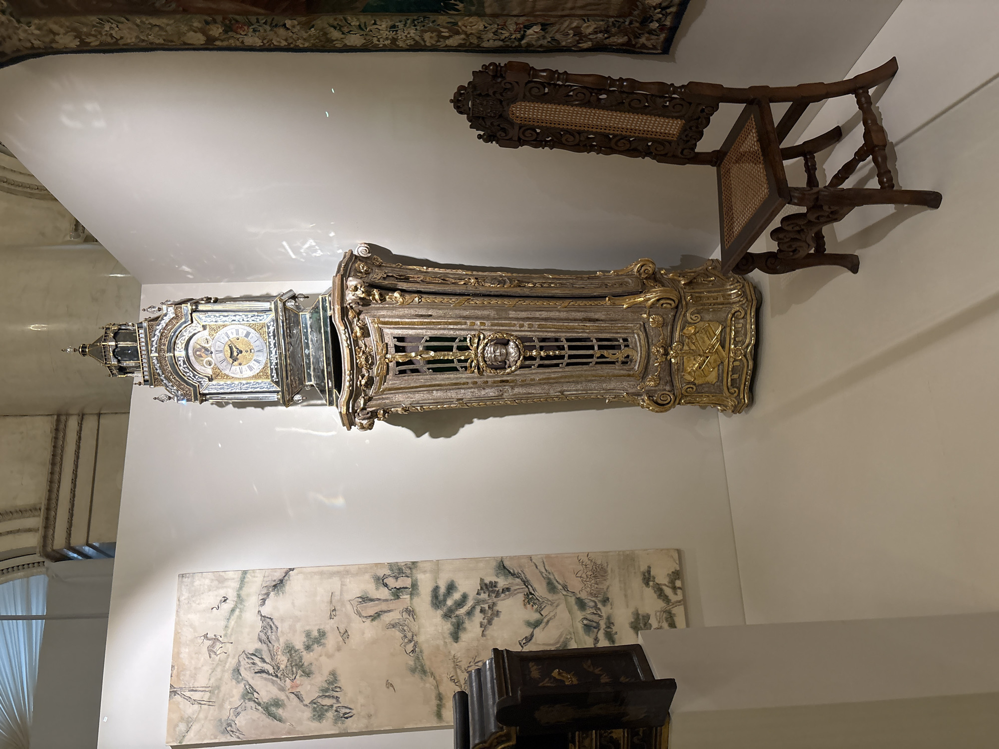 The image shows a room in a building, potentially a museum, in Saint Petersburg, Russia. An ornate grandfather clock with a gold and silver finish stands against a white wall. Above it, a wooden chair with a woven seat leans against the wall. Below, a large painting of a landscape scene hangs. A dark piece of furniture is positioned in front of the painting. A window with curtains is visible to the left. A tapestry is visible at the top of the wall.