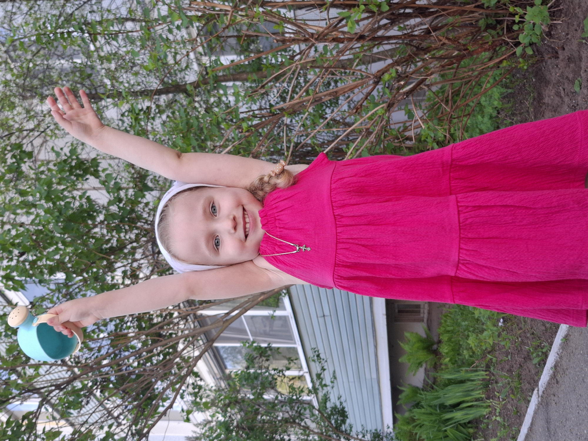A young, blonde-haired girl wearing a pink dress and a white headband is standing outside in Chaikovsky, Russia, with her arms raised. She is holding a blue watering can in her left hand. Behind her are trees with green leaves and a light blue building with white trim. The ground consists of dirt and a concrete sidewalk. The girl is smiling and facing the viewer. A gold necklace with a cross pendant is visible on her chest.