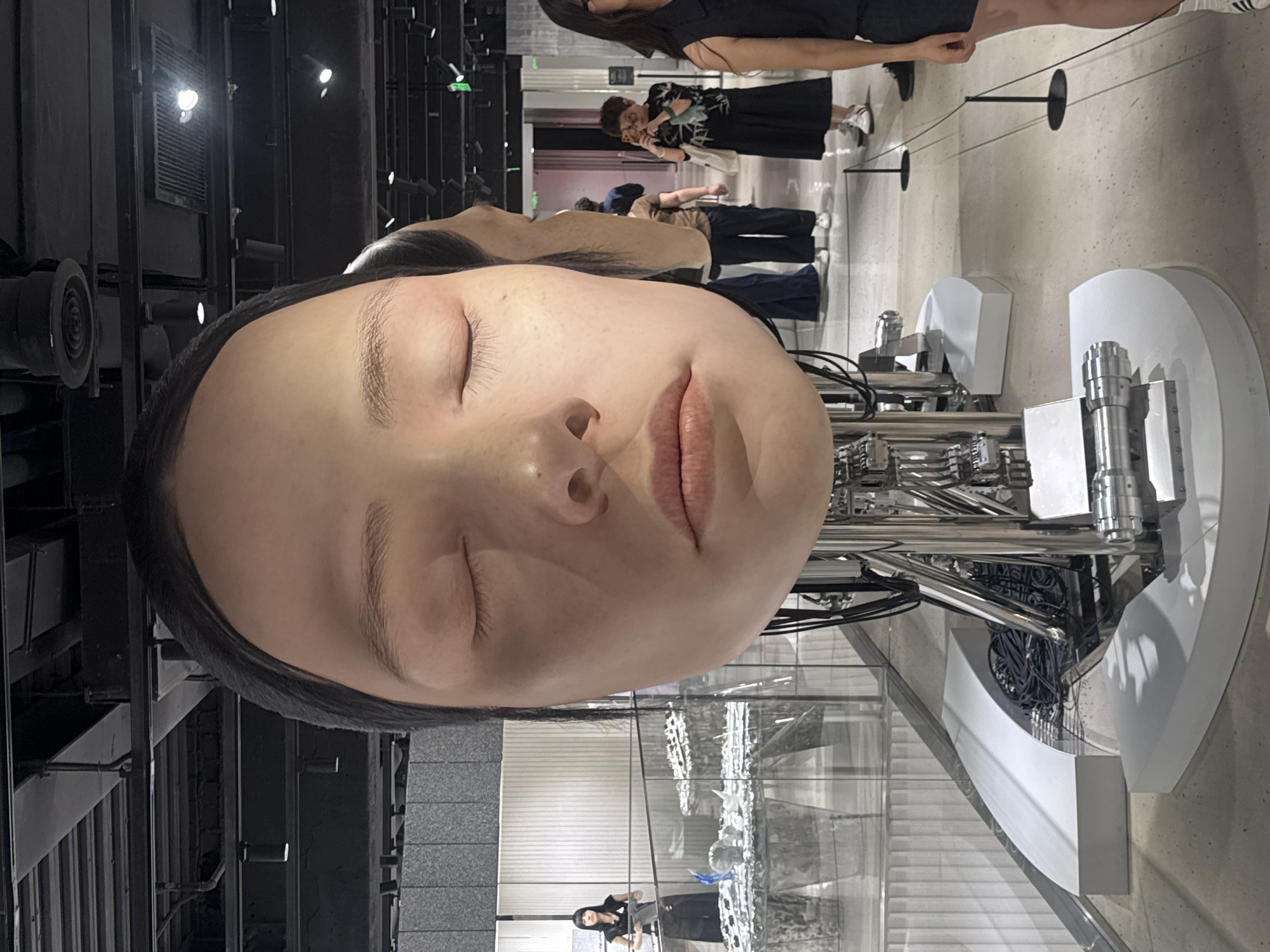The image depicts an art installation featuring a large, hyper-realistic head of an East Asian woman with closed eyes and an expressionless face. The head is mounted on a metallic, mechanical structure, suggesting a robotic or artificial component. In the background, other visitors are visible, some taking pictures, indicating this is a public space, likely a museum or gallery. One woman in the background is seen holding a phone up to take a photo of the installation. To the left of the main sculpture, another similar, albeit smaller, detached head is partially visible. The environment appears to be indoors with industrial-style lighting and overhead structures. The overall impression is one of a contemporary art exhibition. The image was taken in Shanghai, China.