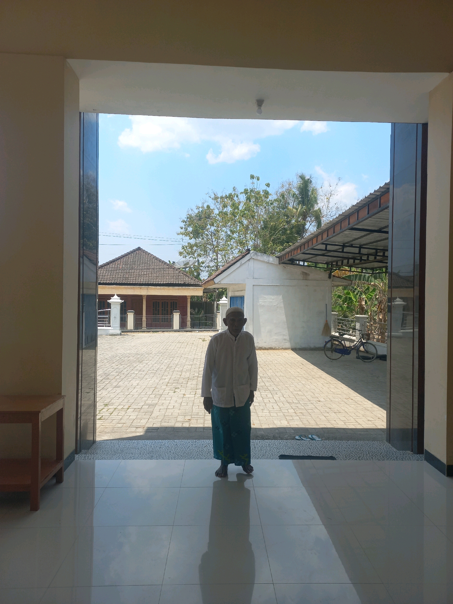 An elderly man wearing a white long-sleeved shirt and a green sarong stands in the doorway of a building, facing outward. He is centered in the frame. The building has a light-colored interior with a tiled floor that reflects the sky and surrounding area. Outside the building, a paved courtyard is visible, with a house featuring a dark tiled roof and a white structure with a corrugated metal roof to the right. A bicycle and a broom are leaning against the white structure. The scene appears to be a residential area in Nguntoronadi, Indonesia, under a clear blue sky with scattered clouds.