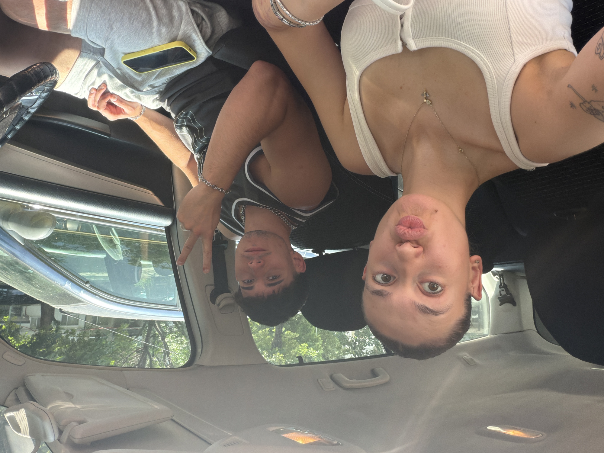 Two individuals are positioned inside a vehicle, with a woman in the front passenger seat capturing a selfie and a man in the driver's seat. The car's interior features a light-colored headliner, overhead lights, and sun visors.
The woman, appearing in the foreground, has light skin, dark hair pulled back, and is wearing a white ribbed tank top. She has a gold chain necklace with a small charm, silver bracelets on her right wrist, and visible tattoos on her left forearm. She is looking directly at the camera with wide eyes and pursed lips.
The man, in the driver's seat, has light skin and dark hair. He is wearing a dark gray tank top with a white design, a silver chain necklace, and a silver bracelet on his left wrist. He is wearing light gray shorts and has a yellow-cased smartphone resting on his lap. His left hand is raised, forming a peace sign, and he is looking towards the camera.
The exterior environment, visible through the vehicle's windows, indicates a bright, sunny day. To the right, a dark-colored sedan is partially visible, appearing parked or moving parallel to the current vehicle. Beyond this, a dense canopy of green trees and parts of a light-colored building with windows are discernible. A thin black wire extends horizontally across the upper background. The scene depicts a typical urban residential setting in Plovdiv, Bulgaria, consistent with a street lined with trees and buildings. No specific landmarks are present to narrow down the precise location within the city.