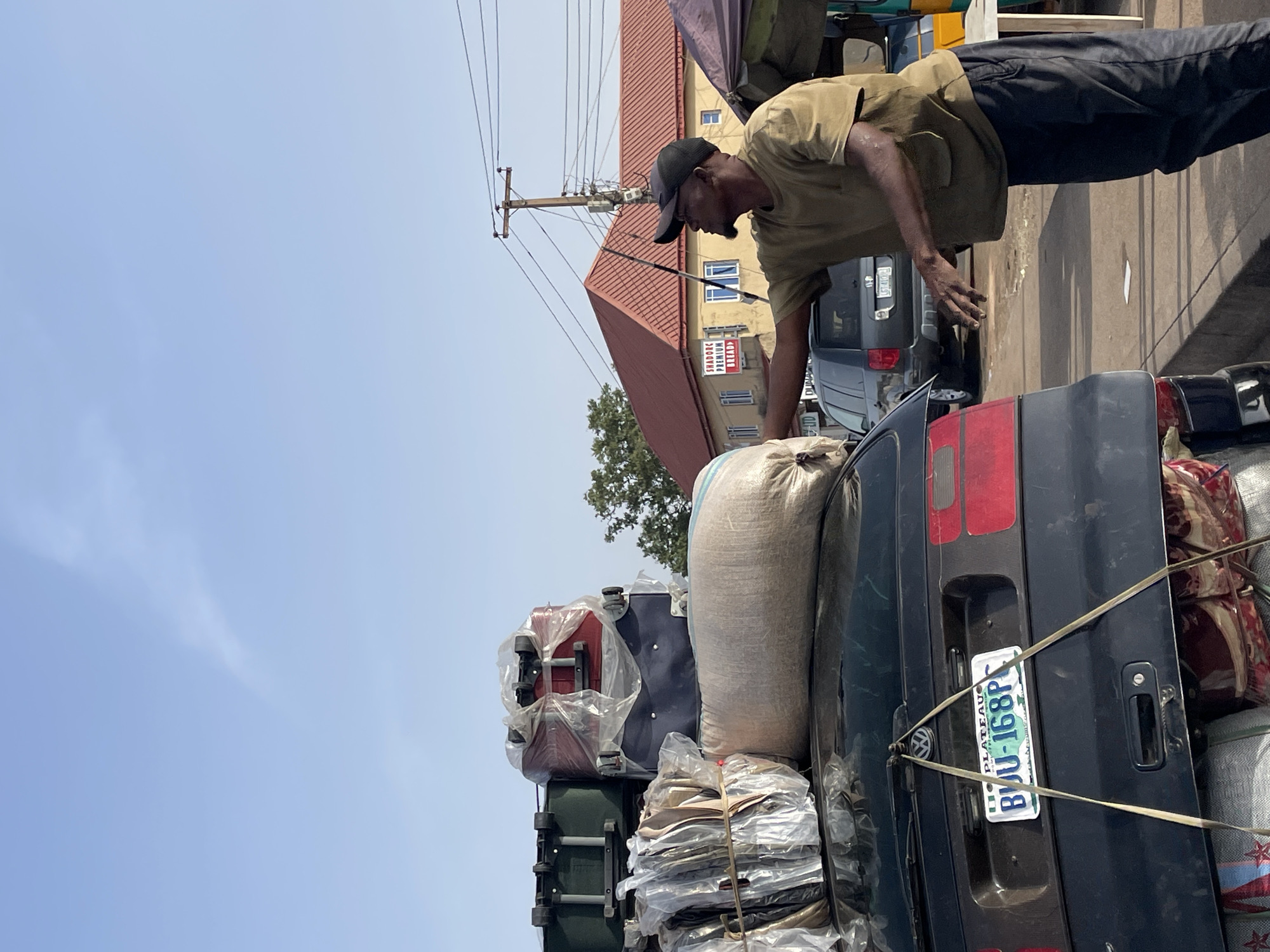 This image captures a bustling street scene in Jos, Nigeria, under a bright, clear daylight sky. The central subjects are a man and a heavily loaded dark-colored station wagon or SUV.
The man, with dark skin, is wearing a khaki short-sleeved shirt, dark trousers, and a black baseball cap. He is leaning over the rear of the vehicle, appearing to be in the process of adjusting or securing the substantial amount of cargo piled on and around the car.
The vehicle is exceptionally heavily loaded with a diverse collection of items. Visible on the roof and rear are a large, light-colored sack (possibly burlap), several pieces of luggage including a red suitcase and a dark-colored one, both wrapped in clear plastic for protection, and various bundles of materials also encased in plastic and tightly secured with straps and ropes. A large, patterned red bag is also visible tied to the side. The vehicle features a Volkswagen emblem on its tailgate and bears a Nigerian license plate that reads "PLATEAU BJU-168PP", indicating registration in Plateau State, Nigeria.
In the background, a multi-story building with a prominent red-tiled roof is visible beneath the blue sky. A sign on this building clearly displays the text "SHADOX PREMIUM BREAD". Numerous overhead power lines stretch across the scene, supported by a wooden utility pole. The overall impression is one of active preparation for transport or movement of goods and belongings, characteristic of a busy urban or semi-urban environment.
