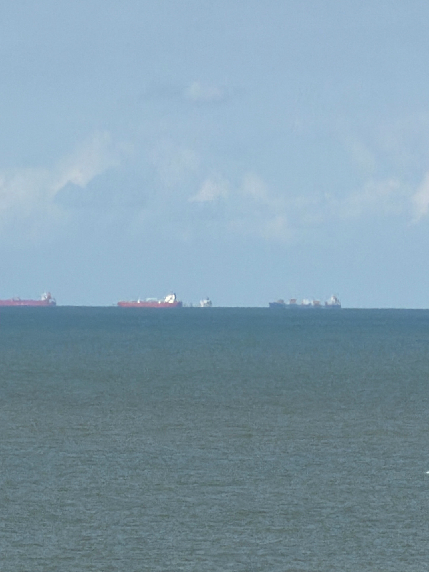 The image shows a wide view of the North Sea off the coast of Ostend, Belgium. Several cargo ships are visible on the horizon. The sea is a deep blue-green, and the sky is a pale blue with scattered white clouds. The overall impression is one of a vast and serene maritime landscape.