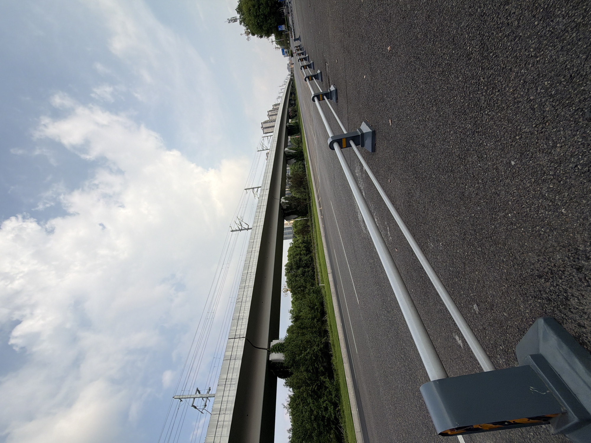 The image shows an empty, multi-lane asphalt road in Jiangjin, China, under a cloudy sky. On the left side of the road, a raised, concrete elevated railway structure runs horizontally across the frame, supported by pillars. Beneath the elevated structure, a dense line of green trees forms a barrier. Along the right side of the road, a series of grey metal safety barriers with yellow reflectors are installed, extending into the distance. The road markings, including white lines, are visible. In the background, modern buildings and telecommunication towers can be seen. The scene is devoid of any visible people or vehicles.