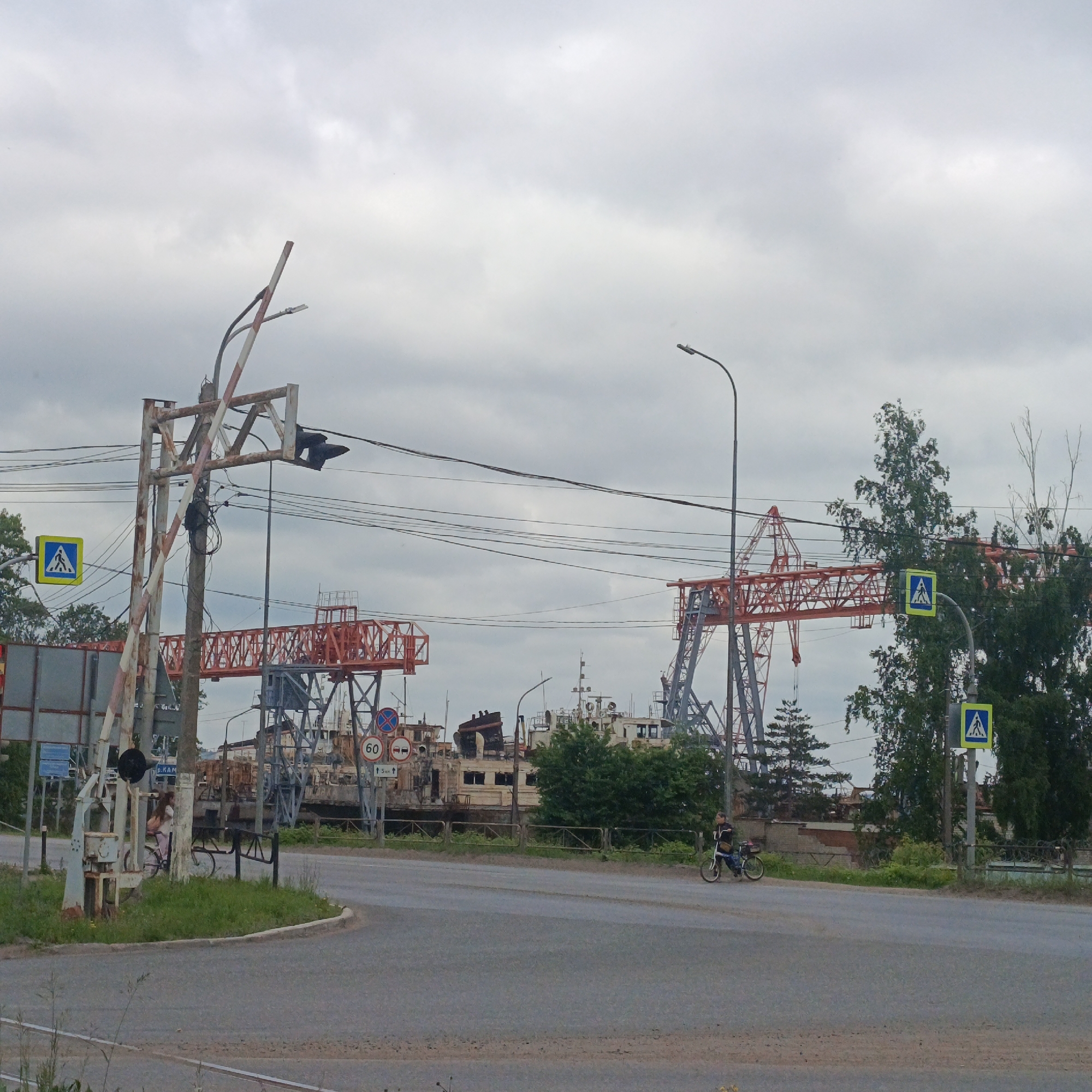 A cloudy, overcast day shot captures a view of a road and a pedestrian crossing near industrial structures. In the foreground, a gray asphalt road curves to the right. A person on a bicycle is visible in the middle ground, riding along the edge of the road.
Further back, an active railroad crossing barrier with red and white stripes is visible. A pedestrian crossing sign, a blue square with a white silhouette of a person walking, is mounted on a pole to the left of the crossing.
The background is dominated by large, industrial cranes with orange lattice structures. Several ships, appearing somewhat derelict or under repair, are docked behind a metal fence. A speed limit sign showing "60" is mounted on a pole, along with a sign indicating "5 Km ↑". The sky is filled with heavy, gray clouds, suggesting a potential for rain. The overall scene depicts an industrial area on a gloomy day.