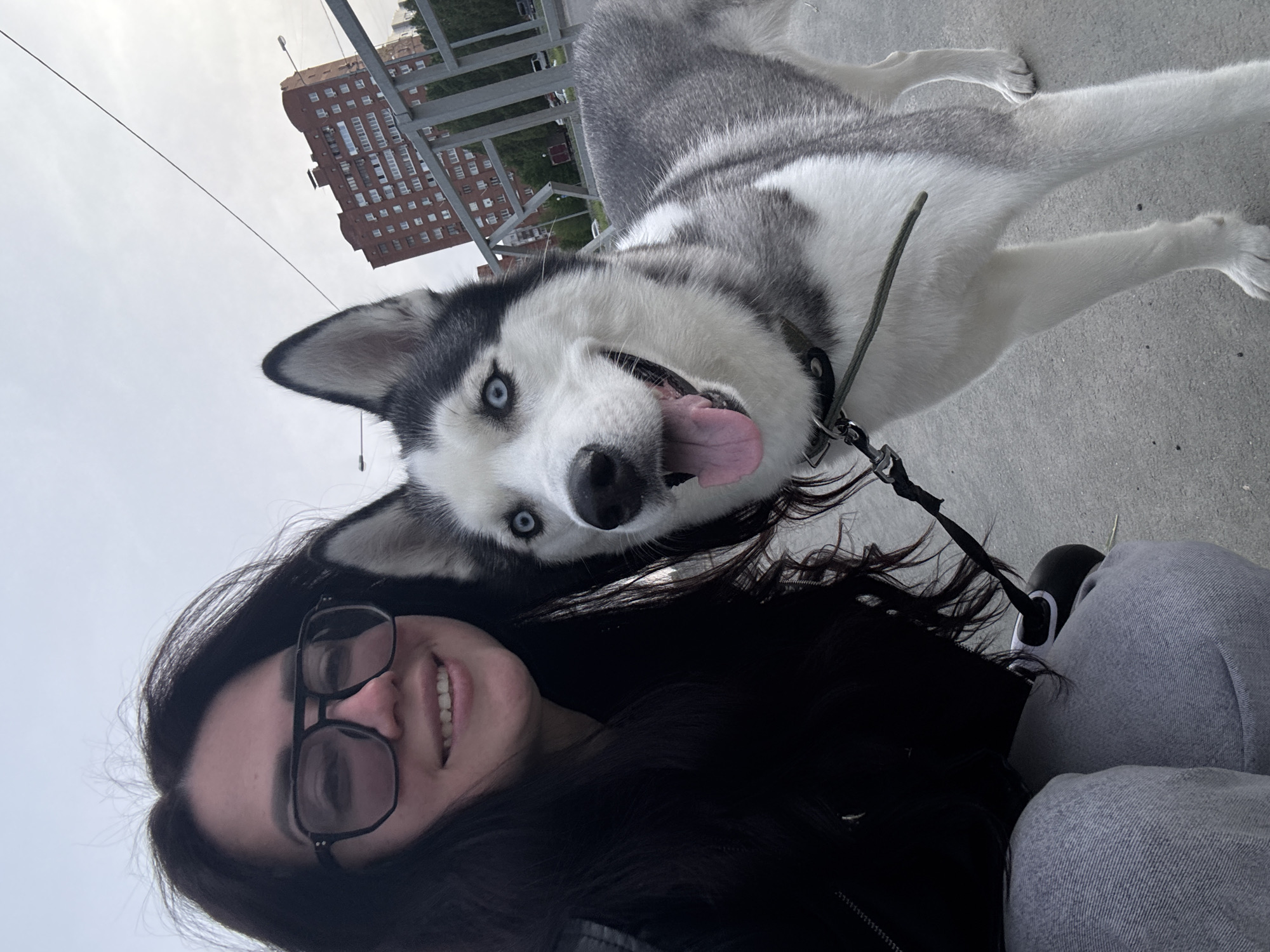 A woman with black, long hair, wearing glasses, is taking a selfie with her husky dog outdoors. The dog is mostly gray and white with piercing blue eyes, panting with its tongue out. They appear to be standing on a paved surface in an urban area, with a tall brick building in the background under a slightly overcast sky. A railing is visible in the background.