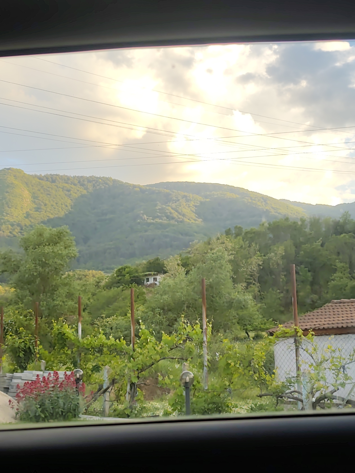The image shows a view of a mountainous landscape under a partly cloudy sky with the sun shining through. In the foreground, there is greenery, including trees and possibly vines. There's also what appears to be a structure with a tiled roof and a fence. The image appears to be taken from inside a vehicle, as evidenced by the window frame visible at the top and bottom of the frame. Power lines are visible stretched across the sky.