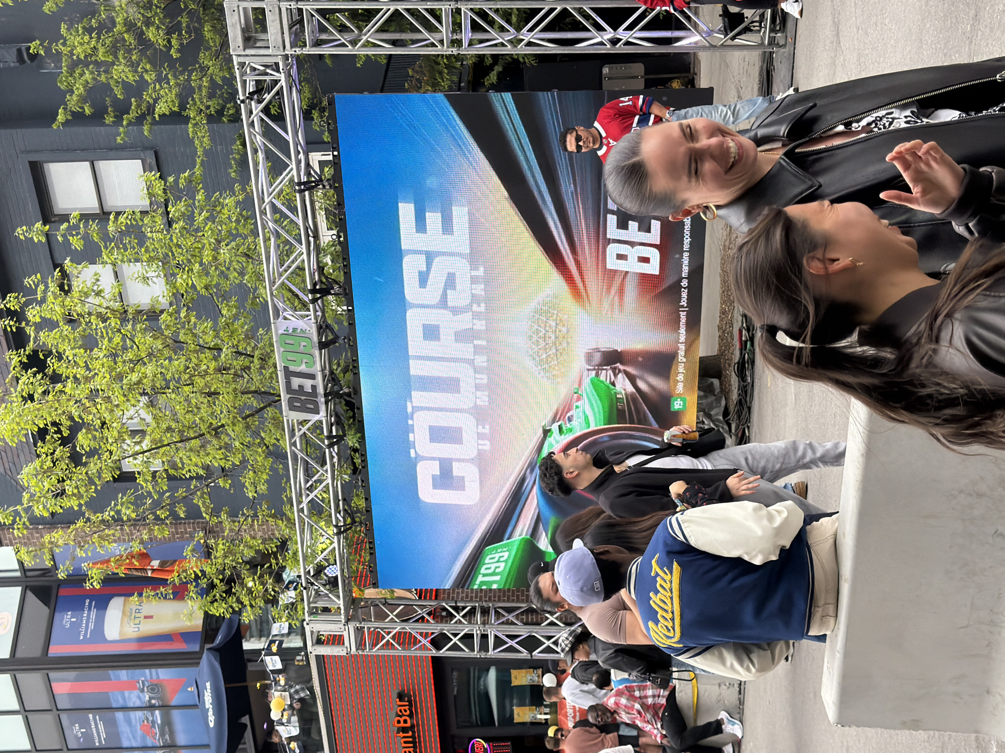 The image captures a scene in Montreal, Canada, likely related to Formula 1 racing. A large video screen displays promotional content for "Course" and "BET99". Two women are visible, smiling and interacting. A crowd of people watches the screen, including some sitting on a concrete structure. The street has signs for "Ultra" and "Williams Racing" and a business named "Ant Bar". There are trees with green leaves in the background.