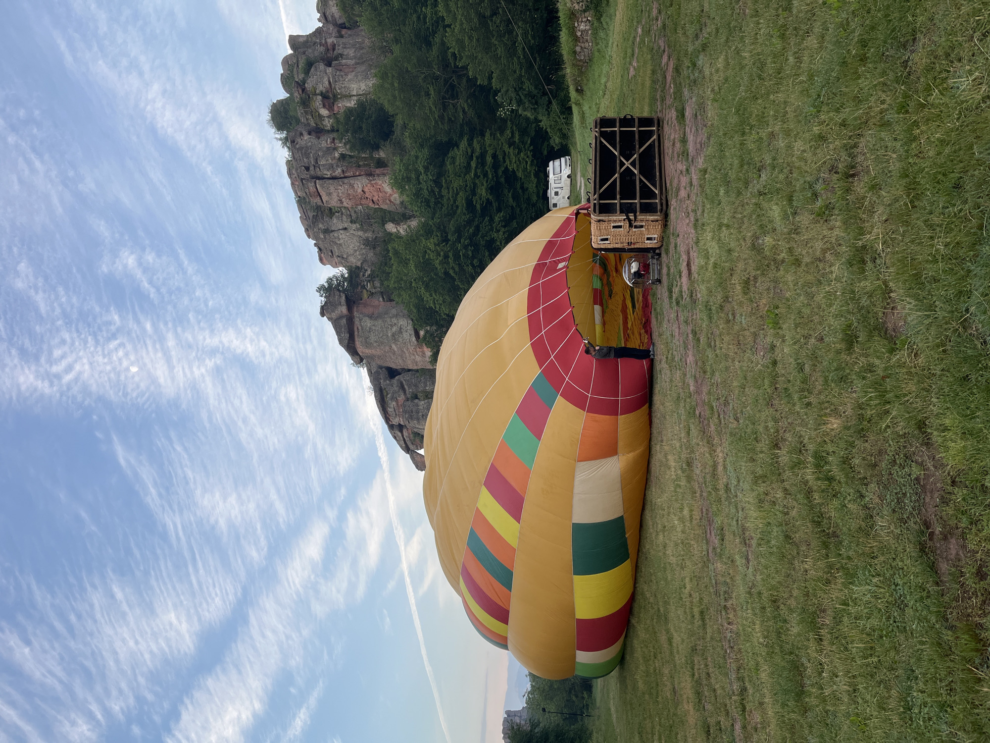The scene captures a hot air balloon activity amidst the Belogradchik Rocks in Belogradchik, Vidin Province, Bulgaria, during what appears to be early morning. A large, deflated or partially inflated hot air balloon, predominantly yellow with vibrant horizontal stripes of red, green, orange, pink, and lighter yellow, lies across a grassy field. Its opening is positioned towards the right foreground.
A single individual, wearing dark trousers and a dark long-sleeved shirt, stands at the mouth of the balloon, facing inward, seemingly engaged in preparing or packing the balloon. Adjacent to the balloon's opening and to the right of the person, a wicker hot air balloon basket lies on its side, revealing its structured base. A silver industrial fan on a trolley is situated near the balloon's opening, between the person and the basket.
The ground consists of green grass with visible dirt paths and some lighter patches of vegetation. In the mid-ground, distinctive reddish-brown rock formations, characteristic of the Belogradchik Rocks, rise dramatically, partially covered by dense green trees and shrubs. Further back on the right, a white camper van is parked amongst the foliage. The sky is a pale blue, traversed by wispy, elongated white cirrus clouds. A faint white crescent moon is discernible in the upper right quadrant of the sky, and a thin white contrail from an aircraft streaks horizontally across the middle-left sky.