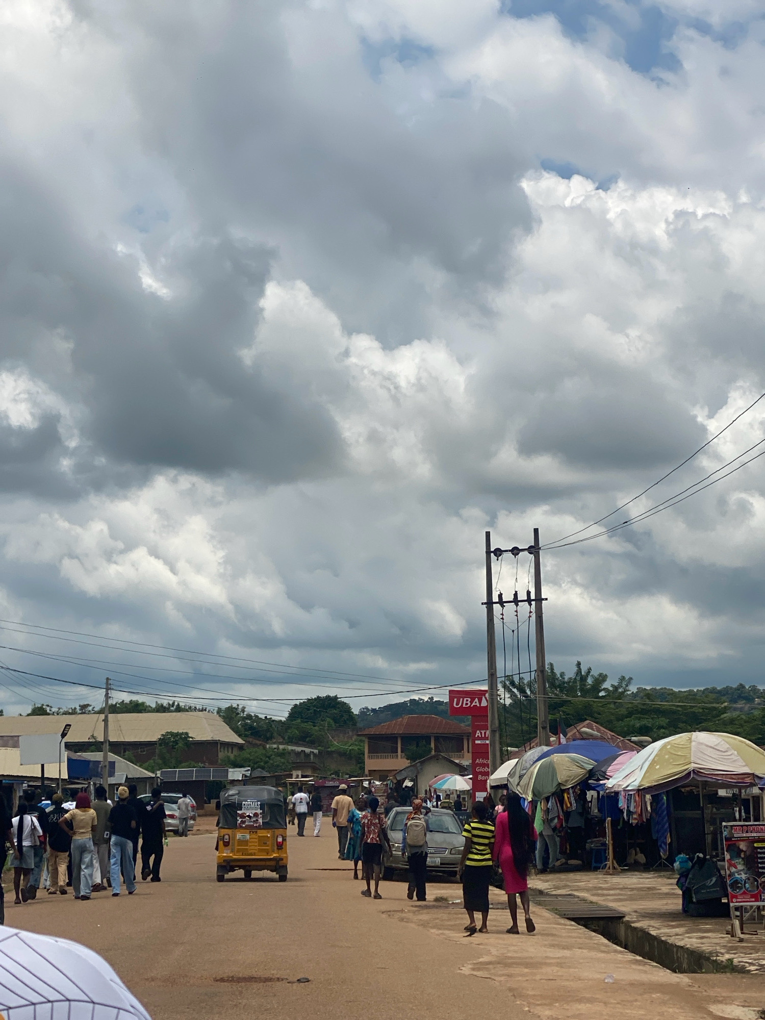 The image depicts a street scene in Iworoko, Nigeria, under a heavily clouded sky. In the foreground, a paved road with a dirt shoulder is visible, with a large group of people walking along the side. Several individuals are dressed casually, and some are carrying bags. To the right, market stalls are set up with colorful umbrellas, displaying clothing and other goods. A yellow three-wheeled vehicle, possibly an auto-rickshaw, is driving away from the camera on the road. In the background, buildings of various sizes and colors are interspersed with trees and vegetation, and a utility pole with wires stands prominently. The overall atmosphere suggests a bustling market or commercial area.