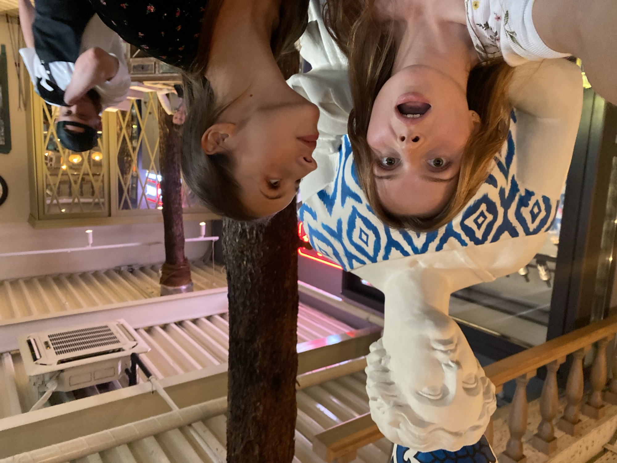 In Solnechnoye, Russia, two young women pose for a selfie with a large, white statue of a woman wearing a blue and white patterned garment and a blue crown. The statue is positioned in the foreground, behind the two women. The woman on the left has her mouth open in a surprised expression and is looking at the camera. The woman on the right is looking upwards, towards the statue's head. Behind them, a tree trunk rises towards a ceiling with exposed beams and an air conditioning unit. Further in the background, several people are visible, including a man wearing a baseball cap who appears to be adjusting his clothing. The setting appears to be an indoor establishment, possibly a restaurant or cafe, with decorative elements and lighting suggesting a public space.