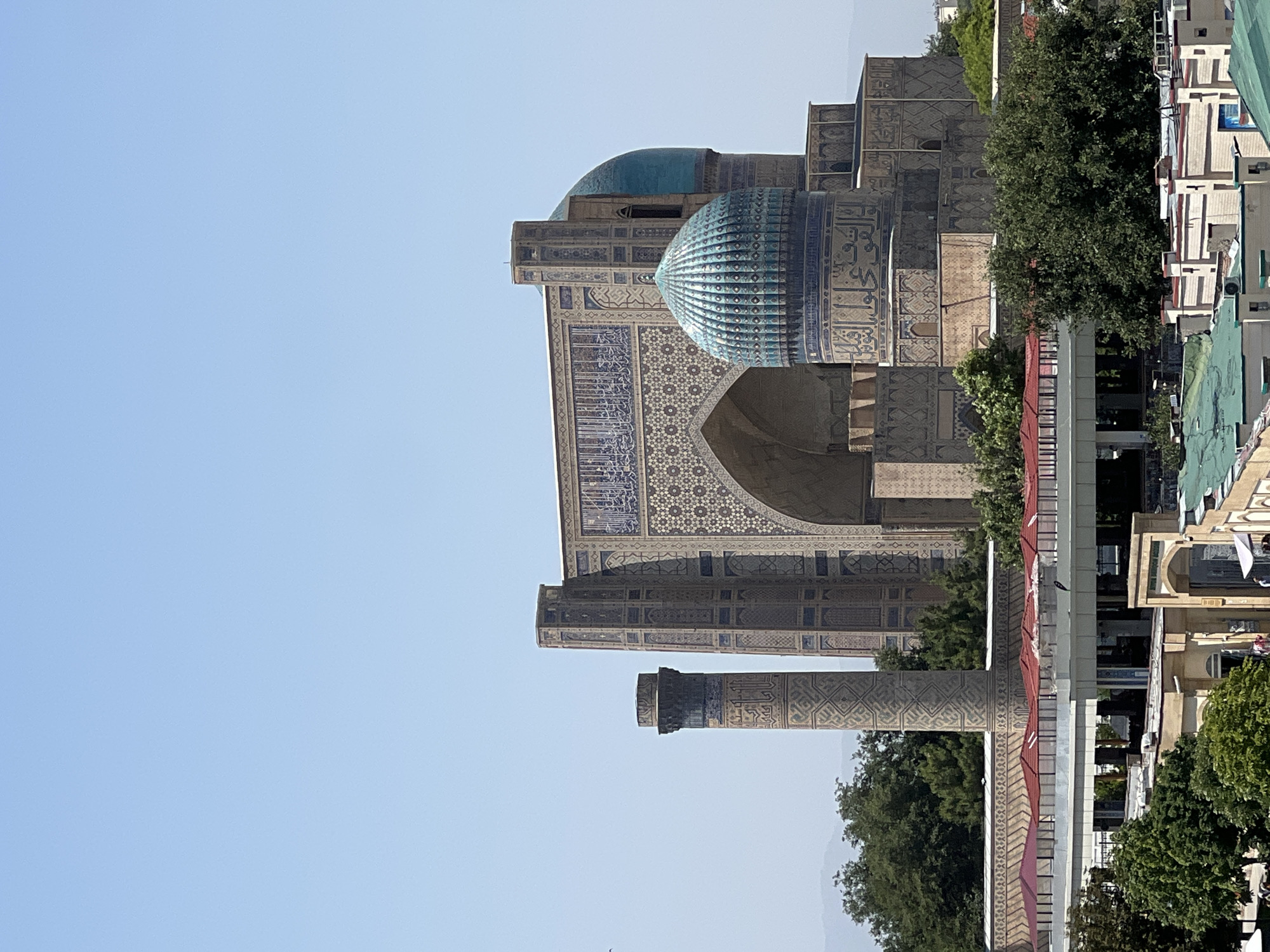 This is a medium-shot, eye-level, outdoor, vertical, daytime photograph of the Bibi-Khanym Mosque in Samarkand, Uzbekistan. The sky is clear and blue. The mosque is the main subject, with its large, ornate architecture dominating the frame. A tall, cylindrical minaret with intricate carvings stands to the left of the main structure, which features a massive arched entrance adorned with blue and white mosaic tiles. Two domes, one a lighter turquoise and the other a darker blue, crown parts of the building. The mosque is surrounded by lush green trees and modern buildings with red-tiled roofs in the foreground, suggesting a bustling urban environment. There are no people visible in the frame. The lighting suggests it is a sunny day.