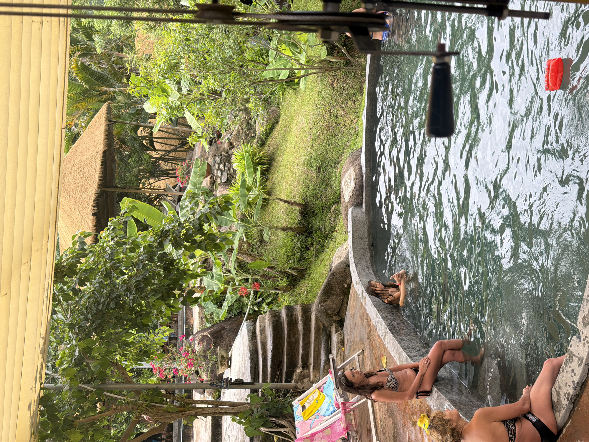 In Khanh Vinh, Vietnam, an outdoor swimming pool area is depicted. The pool is a circular, concrete structure filled with greenish water. Three women are present in the scene. One woman, wearing a leopard-print bikini, sits on the edge of the pool with her legs in the water, looking to her left. Another woman, also in a leopard-print bikini, sits further away on the edge of the pool with her legs in the water, facing towards the pool. A young girl, with wet hair, is partially submerged in the pool, resting her arms on the edge.
Surrounding the pool is lush green vegetation, including trees, banana plants, and grass. A stone staircase with several steps leads up to the left of the pool. A pink and yellow lounge chair is positioned near the staircase. In the background, a thatched-roof pavilion is visible amongst the trees. The sky is partially obscured by a yellow canopy. A red floatable object is seen in the water, near the foreground. The overall impression is a natural, tropical resort setting.