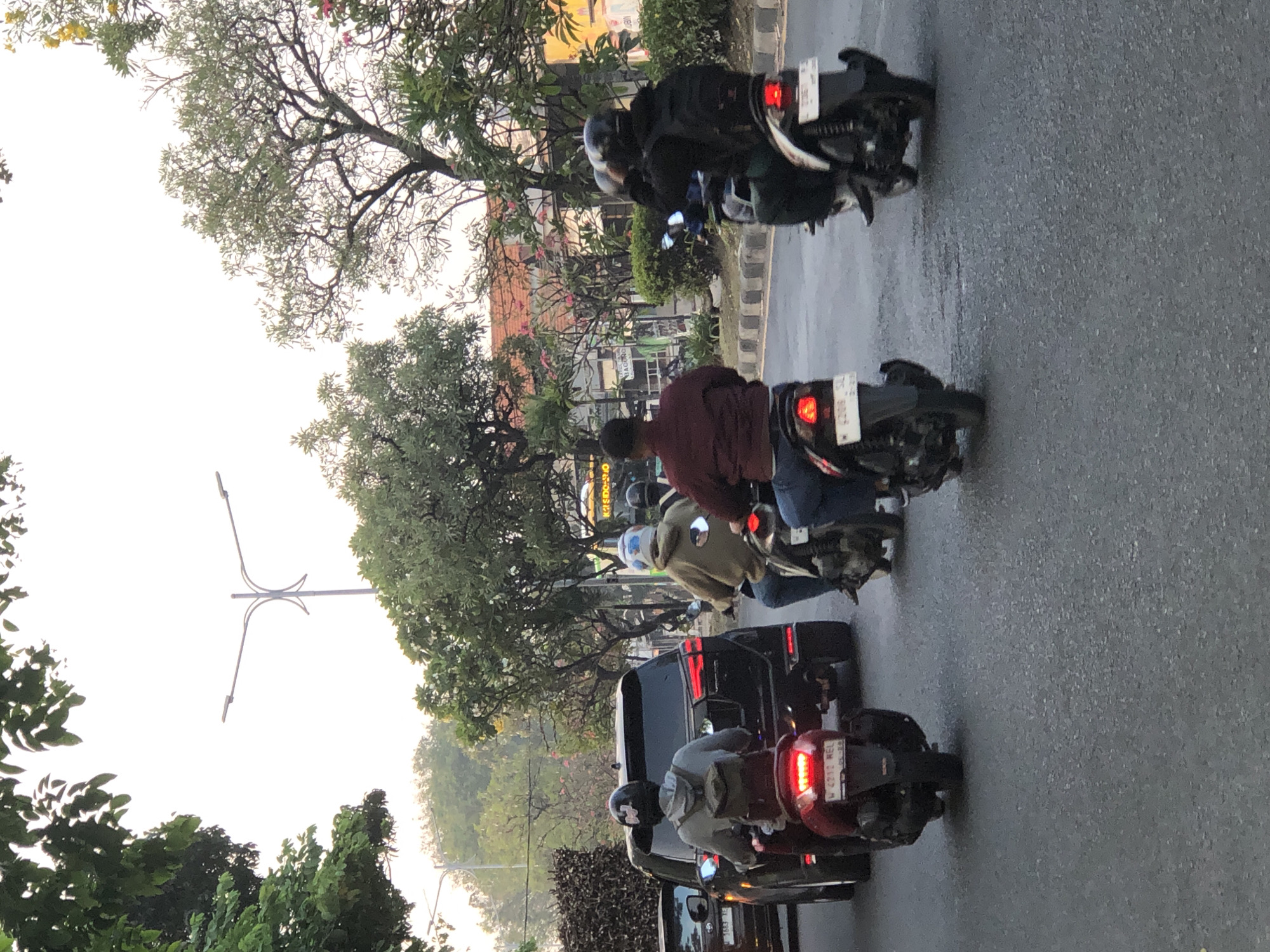 The image depicts a street scene in Ngampelsari, Indonesia, likely during the late afternoon due to the soft, diffused lighting. The foreground is dominated by a paved road with visible tire marks and some minor surface imperfections. Several motorbikes and a dark SUV are in motion, suggesting traffic.
On the left, a dark SUV is the primary subject, with its rear visible. A red motorbike is positioned in front of the SUV, with its license plate reading "W 4913 HEL." The rider of this motorbike is wearing a grey hoodie and a helmet with a black sticker.
Further into the scene, a motorbike ridden by an individual in a tan hoodie is in the middle lane, facing away from the viewer. To the right of this motorbike, another motorbike is occupied by two individuals. The rider is wearing a dark jacket and a backpack, while the passenger is also wearing dark clothing and a helmet. A fourth motorbike, ridden by someone in a maroon jacket, is visible on the right side of the road.
The background features lush green trees, some with pink flowers, and buildings with orange tiled roofs. A streetlamp with two lights is positioned in the middle ground. The overall environment suggests a moderately busy urban or suburban street in Indonesia.