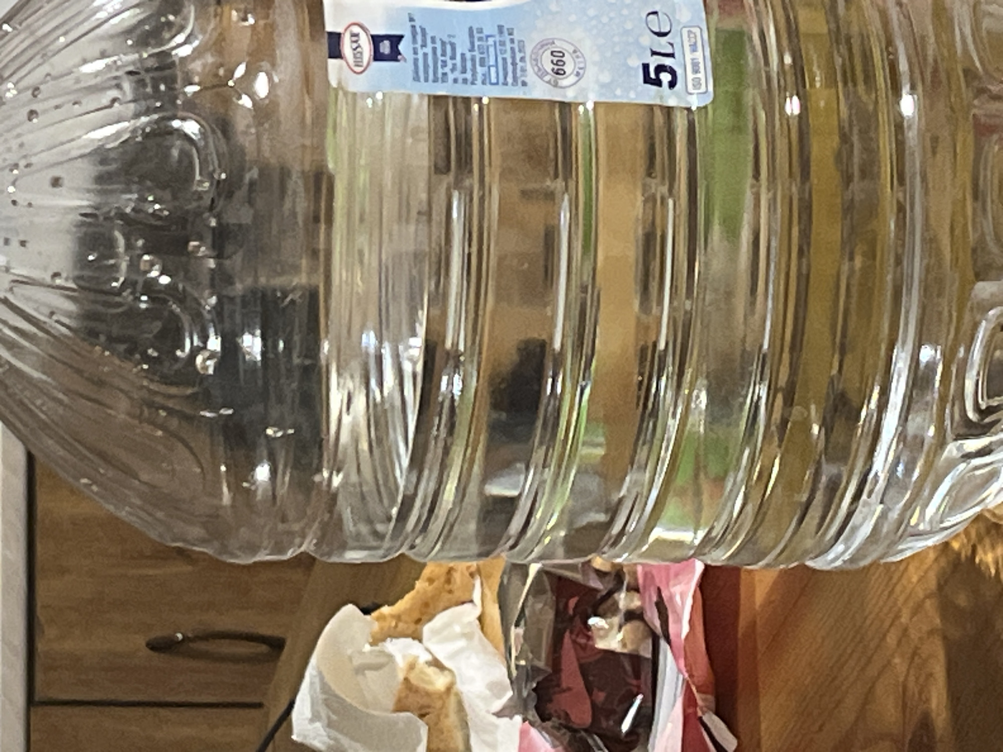 A close-up shot focuses on a 5-liter plastic bottle of Hissar brand mineral water. The bottle is partially filled and has a label indicating a capacity of 5 liters and a number "660". Beneath the bottle, there are remnants of food packaging, including what appears to be discarded pastries in white paper and a candy wrapper. The surface beneath them is made of wood, possibly a table or countertop. A drawer with a metal handle is visible in the lower-left corner of the frame. The location can't be identified without additional information.