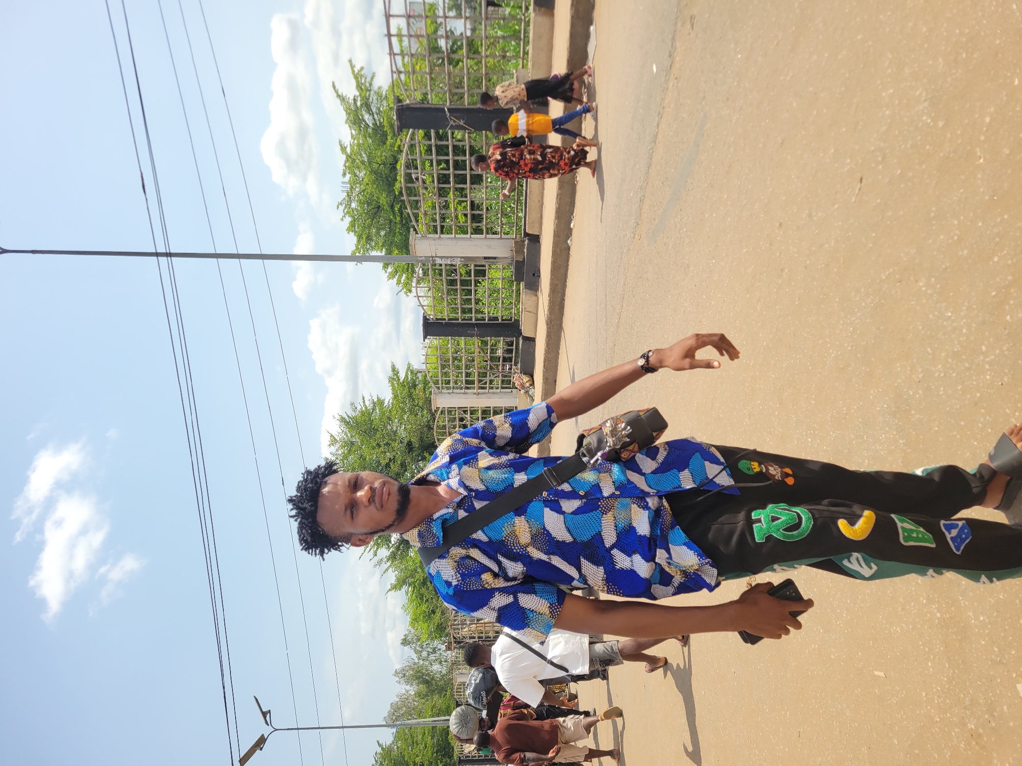 The image captures a vibrant street scene in Onitsha, Nigeria, during bright daylight. The sky is clear with scattered white clouds, and multiple utility lines crisscross the upper part of the frame.
The central subject is a young Black man, looking directly at the camera with a calm expression. He has short, dark, curly hair and a neat beard. He is wearing a long-sleeved, collared shirt featuring a bold geometric pattern in shades of blue, white, and yellow. A black shoulder bag with a patterned flap is slung across his body. In his left hand, he holds a dark smartphone. He wears black pants adorned with several large, colorful patches, including a green letter, a yellow crescent shape, and blue and green abstract designs, along with some cartoonish figures. He is wearing grey slide sandals.
In the mid-ground and background, several other people are visible. Some individuals are walking past a tall black metal fence or barrier, which suggests a public walkway, bridge, or divider alongside the street. Green trees and foliage are visible beyond this structure. The ground is a light brown paved surface, indicative of a road or pedestrian area. A utility pole with a street light extends upwards on the left side of the image. The overall setting is an active urban environment.