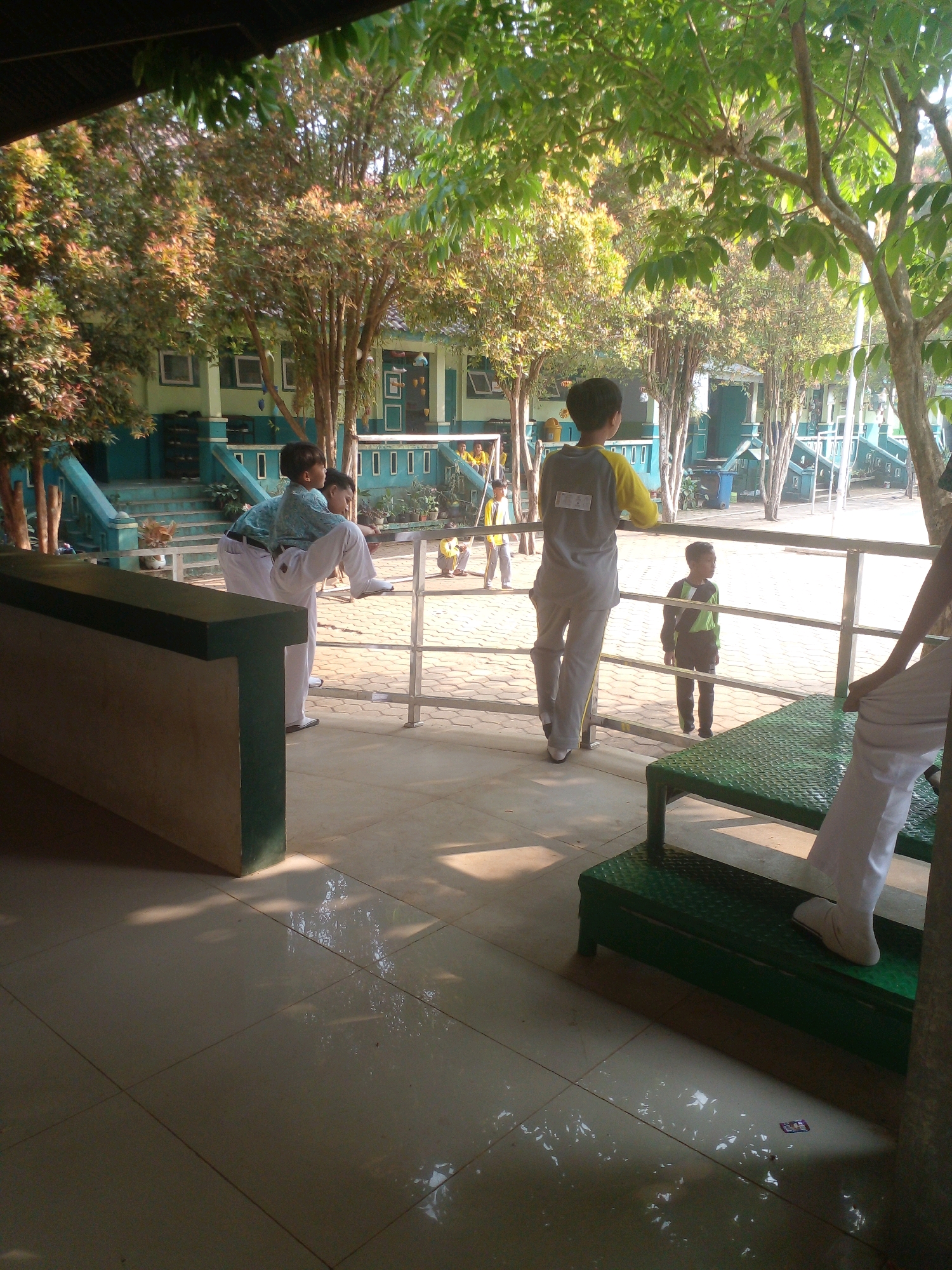 The image depicts an outdoor schoolyard scene in Banyuputih, Indonesia. In the foreground, a green metal bench or platform is visible with a person's legs and feet wearing white pants and shoes. Behind this, a group of school-aged children are present. Two boys in school uniforms are standing near a metal railing. One boy is wearing a blue patterned shirt and white pants, while the other is wearing a grey shirt with yellow accents and grey pants. Another young boy in a dark shirt with green accents and dark pants stands further away. In the background, a school building with green and blue accents is visible, surrounded by trees. The ground is paved with hexagonal tiles. Sunlight casts shadows across the scene, indicating it is daytime.