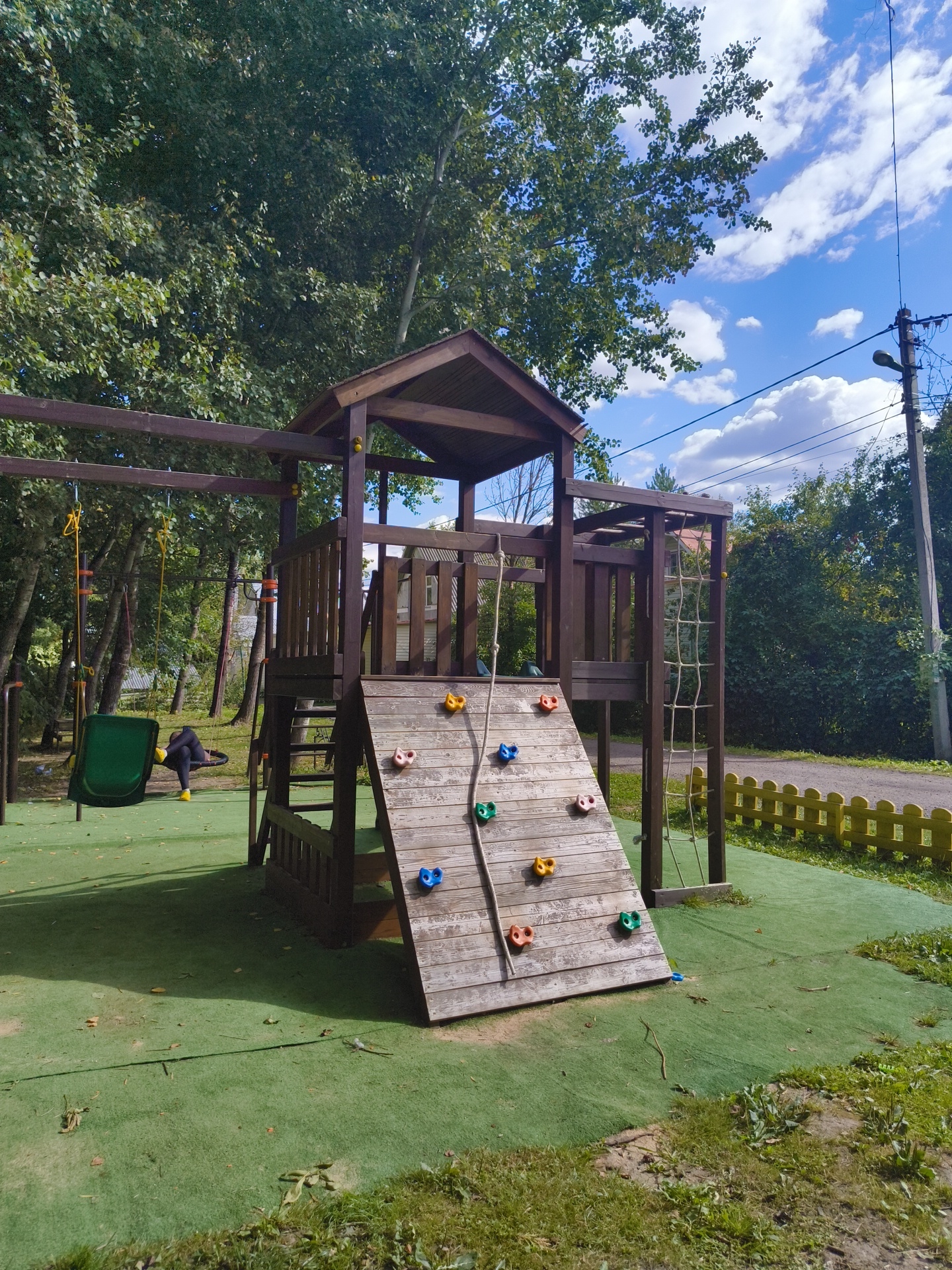A wooden playground structure with a climbing wall, slide, and swings is situated on a green artificial turf surface. The structure features a small wooden house with a roof, wooden railings, and a wooden ladder. The climbing wall is made of wooden planks and has colorful climbing holds attached. A green swing with a person sitting on it is visible to the left of the structure. In the background, trees, a utility pole with wires, a short yellow fence, and a dirt road are present. The sky is blue with scattered white clouds. The location is identified as SNT Pravdist-2, Russia.