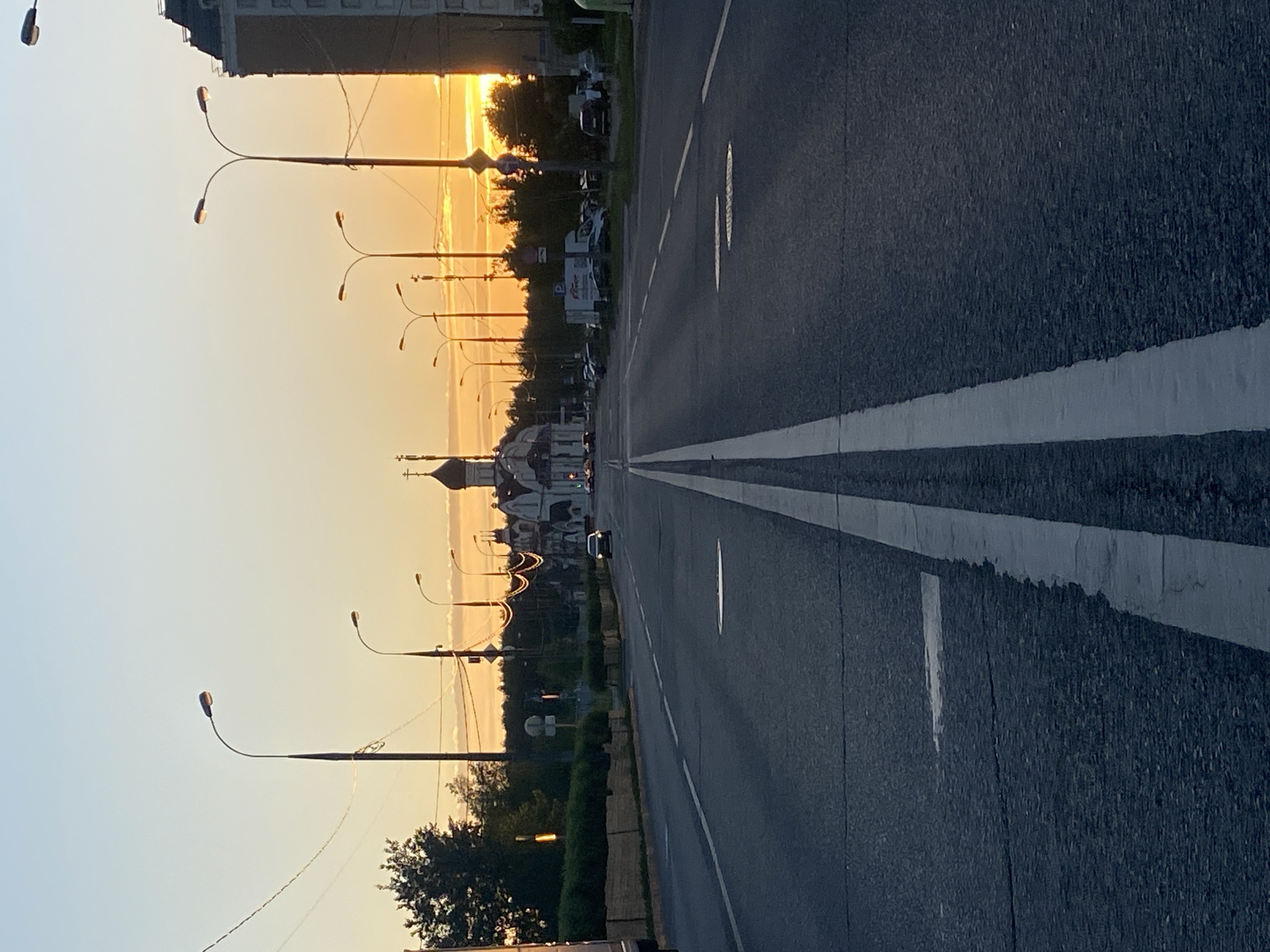 A road in Moscow, Russia, is depicted during sunset. The road has two lanes marked by white lines. Streetlights line the left side of the road. A church with a steeple is visible in the distance. Buildings and trees border the road, with a building in the background. A car is on the road. The sky is yellow and orange from the setting sun.